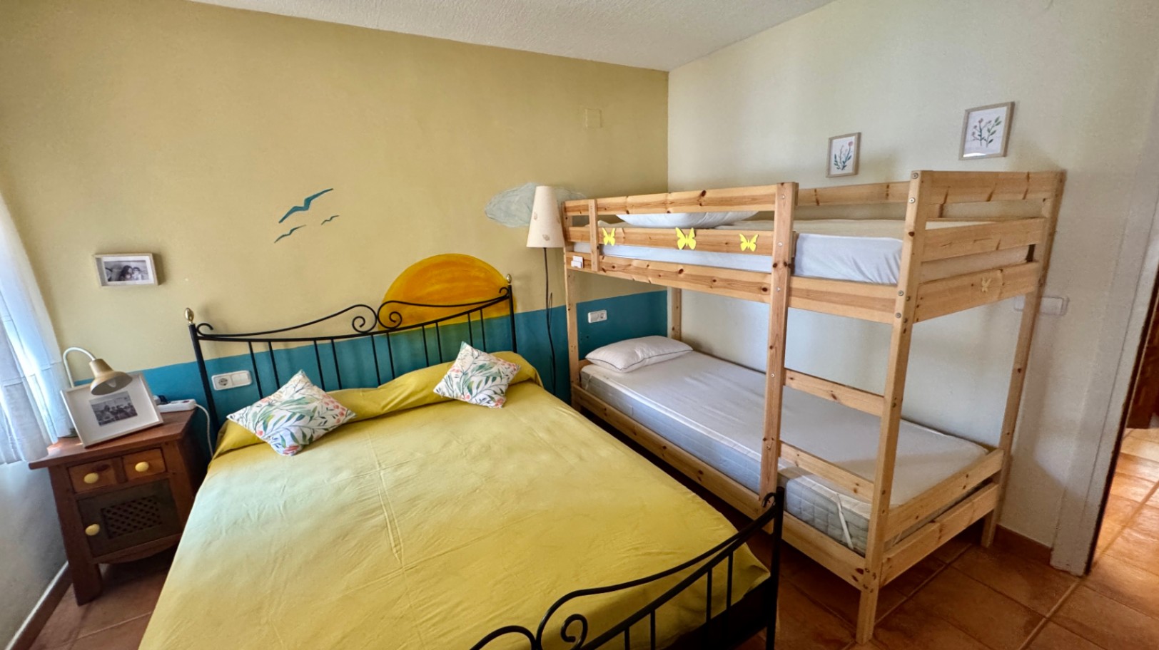 Schlafzimmer mit einem Doppelbett aus schwarzem Schmiedeeisen und einem Etagenbett aus Naturholz. Mit Terrakotta-Fliesenboden und einer dekorativen Wandmalerei.