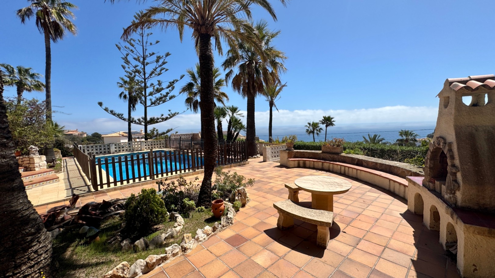 Terracotta-Terrasse mit Steinsitzgruppe, rustikalem Außenkamin und umzäuntem Pool mit Blick auf das Meer und Palmen.