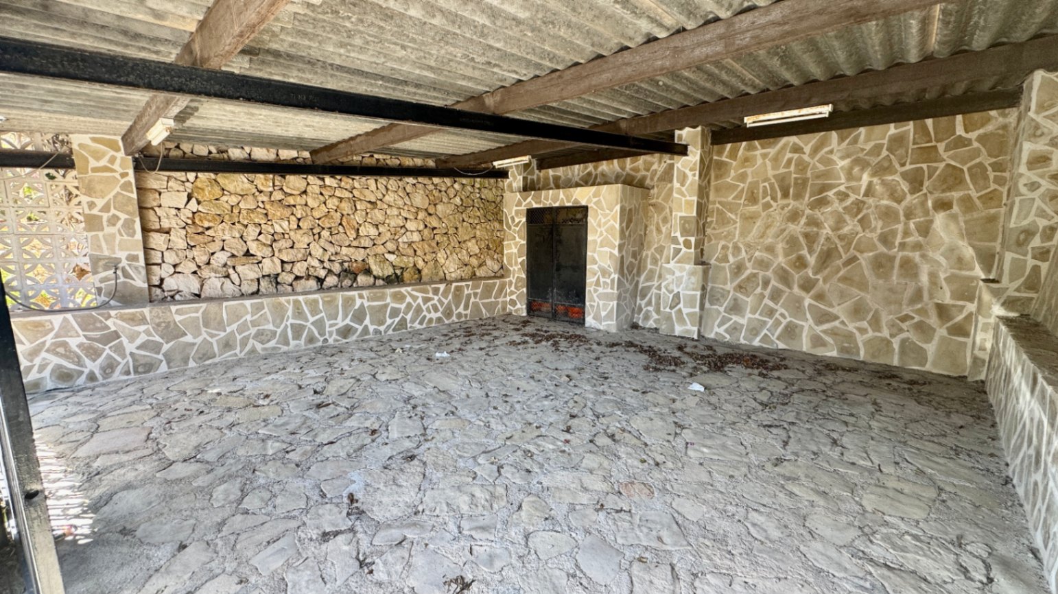Überdachter Außenbereich mit rustikalen Steinwänden und -böden, Holz- und Metallbalken sowie einem Wellblechdach.