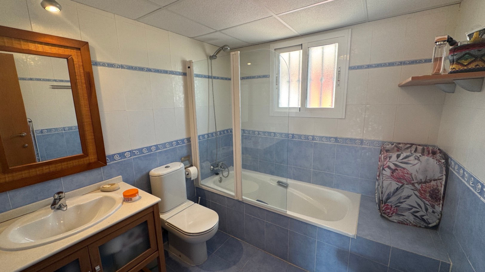 Badezimmer mit blau-weißen Fliesen, einer Badewanne mit Glasabtrennung, einem Holzrahmen-Spiegel und einem passenden Waschtischunterschrank.