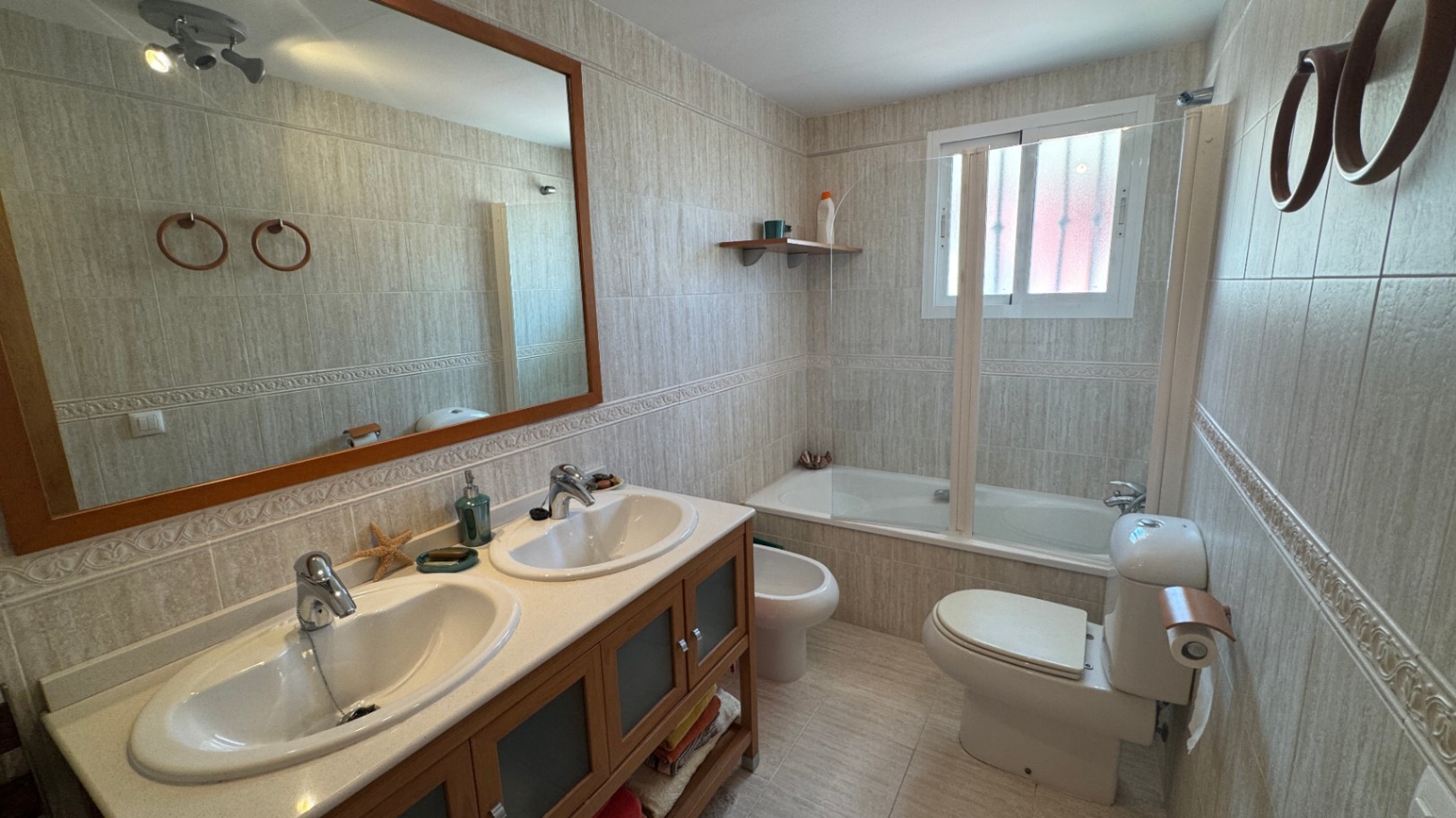 Badezimmer mit Doppelwaschtisch auf einem Holzunterschrank, einer Badewanne mit Glasabtrennung und hellen Wandfliesen mit dekorativer Bordüre.