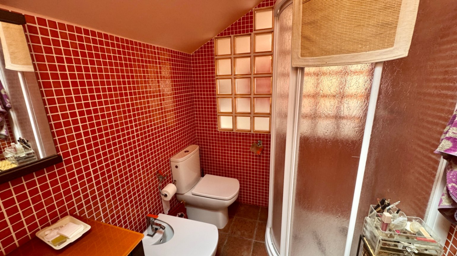 Badezimmer mit roten Mosaikfliesen, Dachschräge und einer geschwungenen Eckdusche. Ausgestattet mit WC, Bidet und einem Glasbausteinfenster für Tageslicht.