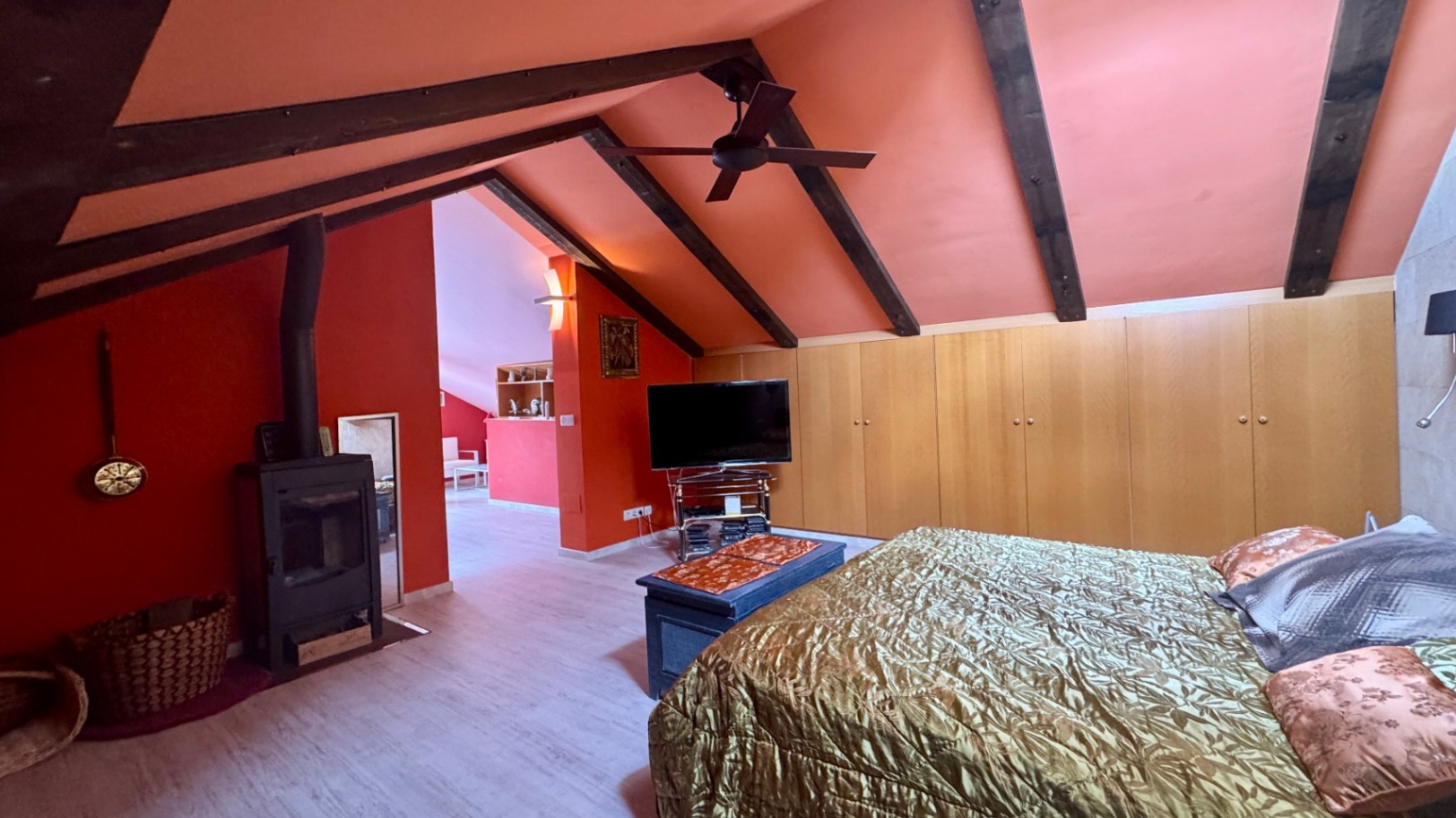 Dachgeschoss-Schlafzimmer mit dunklen Sichtbalken, terracottafarbenen Wänden, einem Kaminofen und maßgefertigten Einbauschränken aus Holz.
