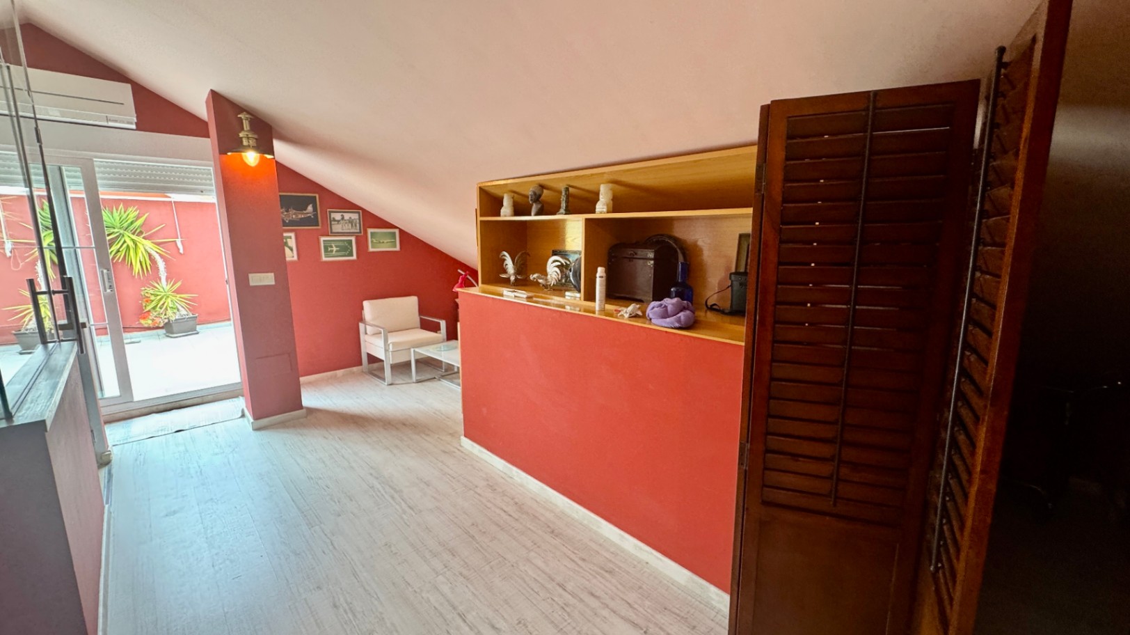 Dachgeschosszimmer mit terrakottafarbenen Wänden, Schrägen und integrierten Holzregalen. Eine Glastür führt zur privaten Terrasse mit mediterranen Topfpflanzen.