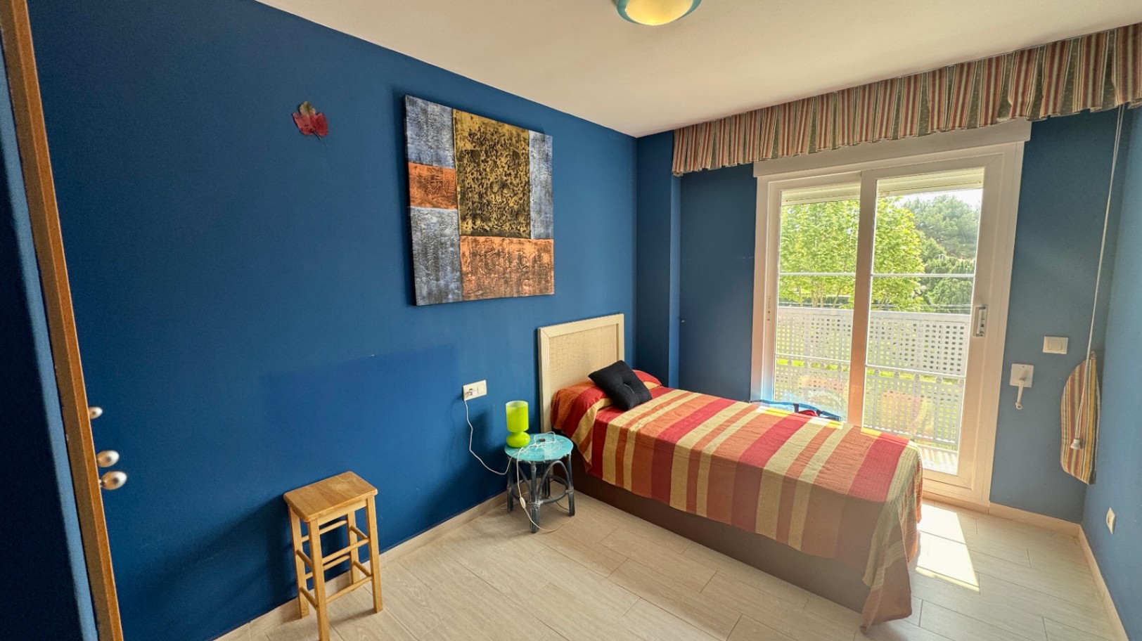 Einzelzimmer mit tiefblauen Wänden, einem Kopfteil aus Korbgeflecht und einer großen Glasschiebetür, die auf einen privaten Balkon mit Blick ins Grüne führt.