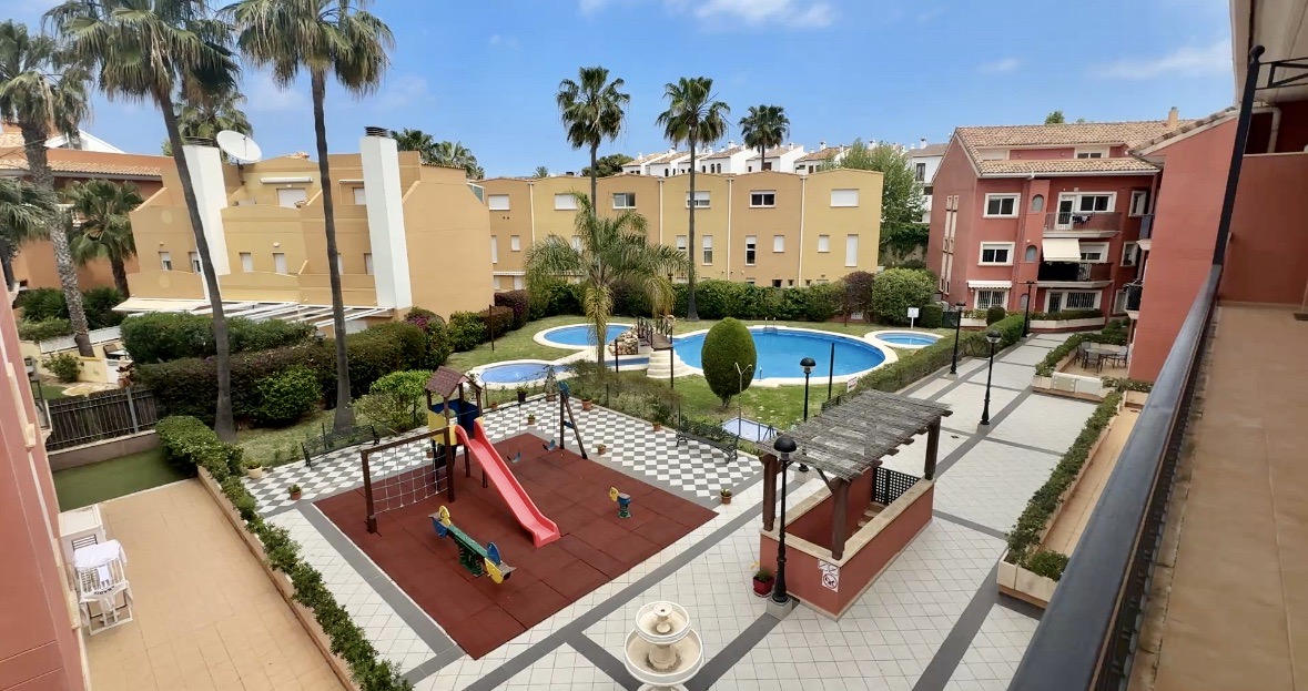 Erhöhter Blick auf den Gemeinschaftsbereich mit einem mehrteiligen Pool, einem Kinderspielplatz mit Fallschutzboden und gepflegten Gartenanlagen mit Palmen.