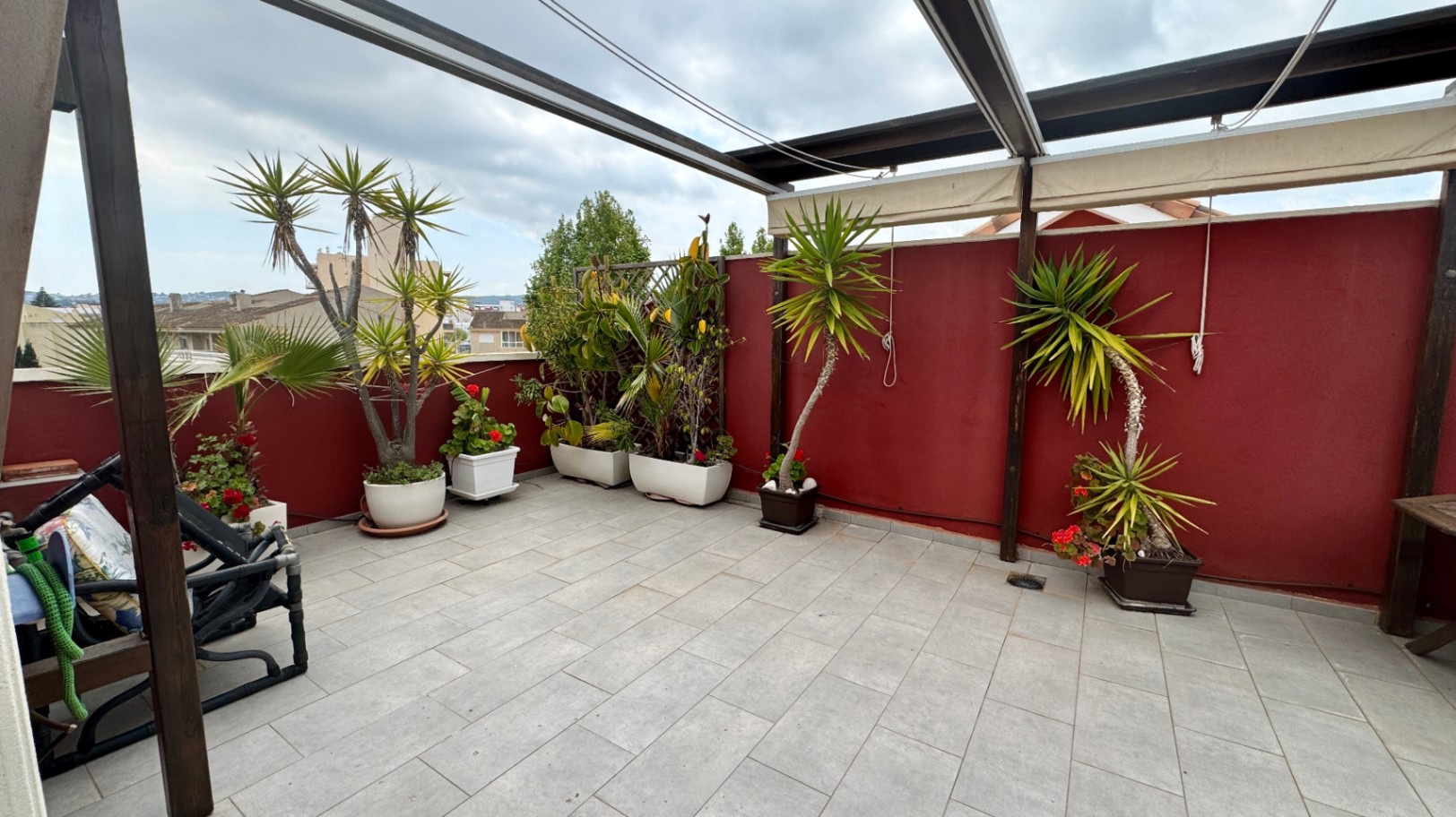 Großzügige Dachterrasse mit grauen Bodenfliesen, einer Holzpergola mit Markisen und verschiedenen Kübelpflanzen vor einer markanten weinroten Wand.