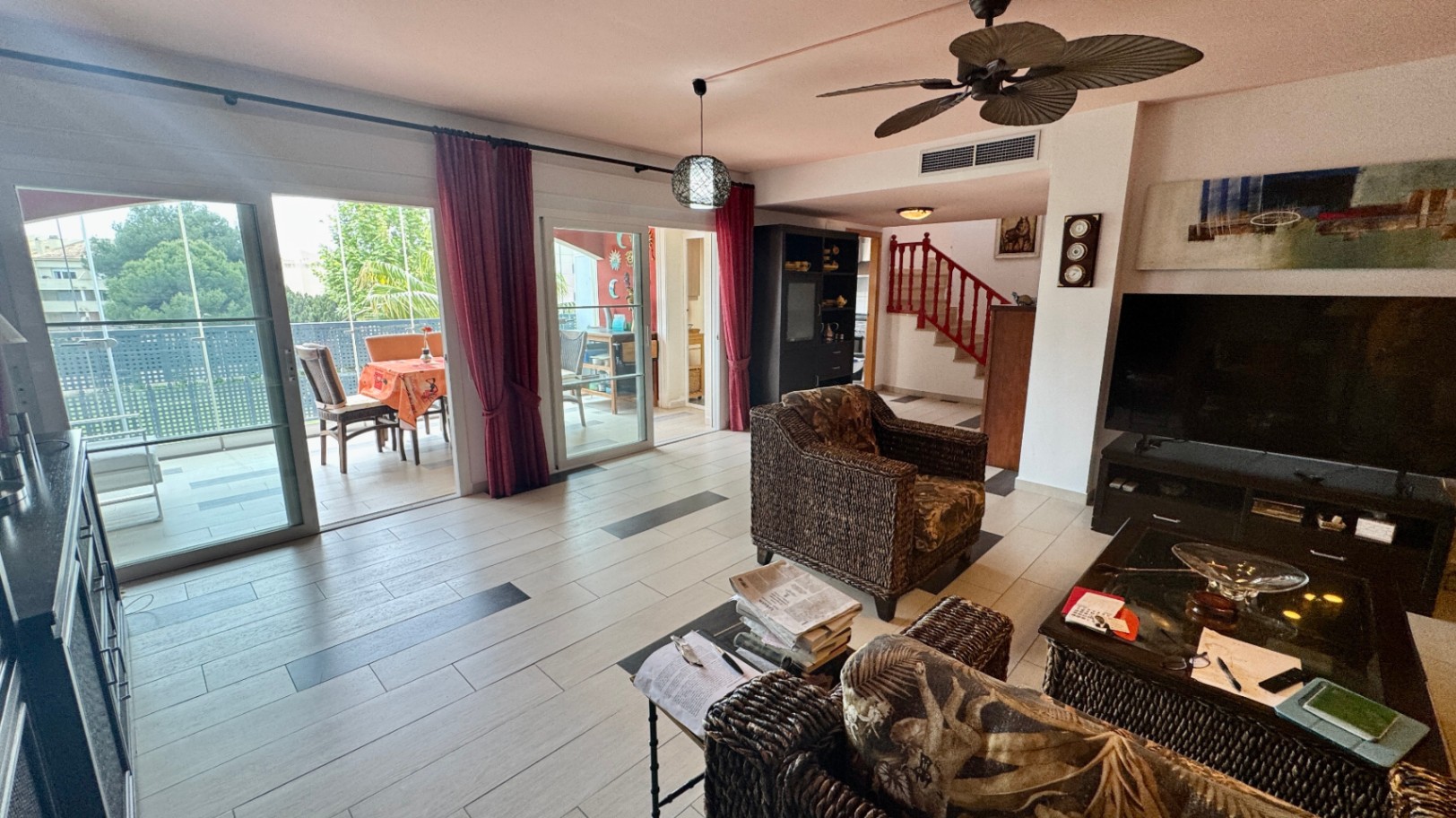 Heller Wohnbereich mit Rattansesseln, Fliesenboden mit dunklen Akzenten und Glasschiebetüren zur Terrasse. Ein Deckenventilator und eine rote Holztreppe ergänzen das Interieur.