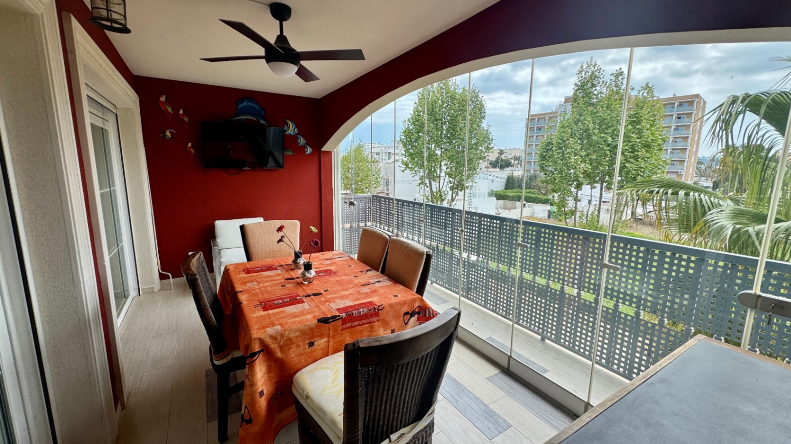Verglaste Terrasse mit Glasschiebewänden, roter Akzentwand mit Fernseher und Essbereich mit Blick auf die umliegenden Bäume und Gebäude.