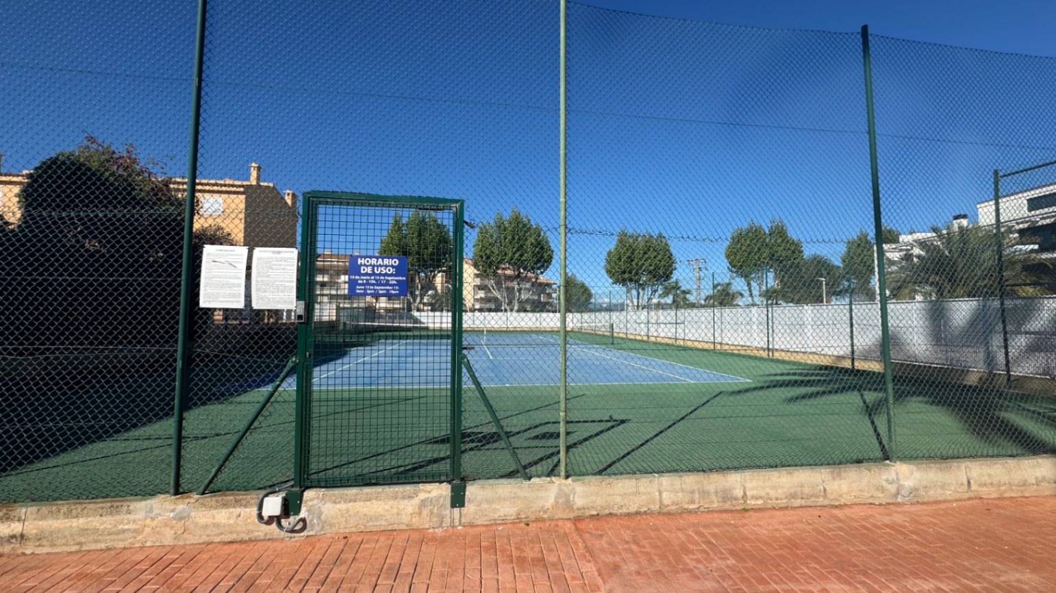 Außen-Tennisplatz mit blau-grünem Hartplatzbelag, umgeben von einem hohen Maschendrahtzaun und einem abschließbaren Metalltor für den Zugang.
