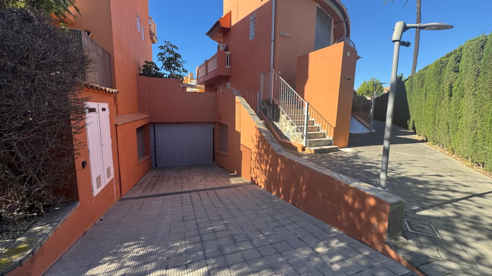 Außenansicht mit gepflasterter Einfahrt zur Privatgarage, terrakottafarbener Fassade und Außentreppe mit Metallgeländer neben einer hohen Hecke.