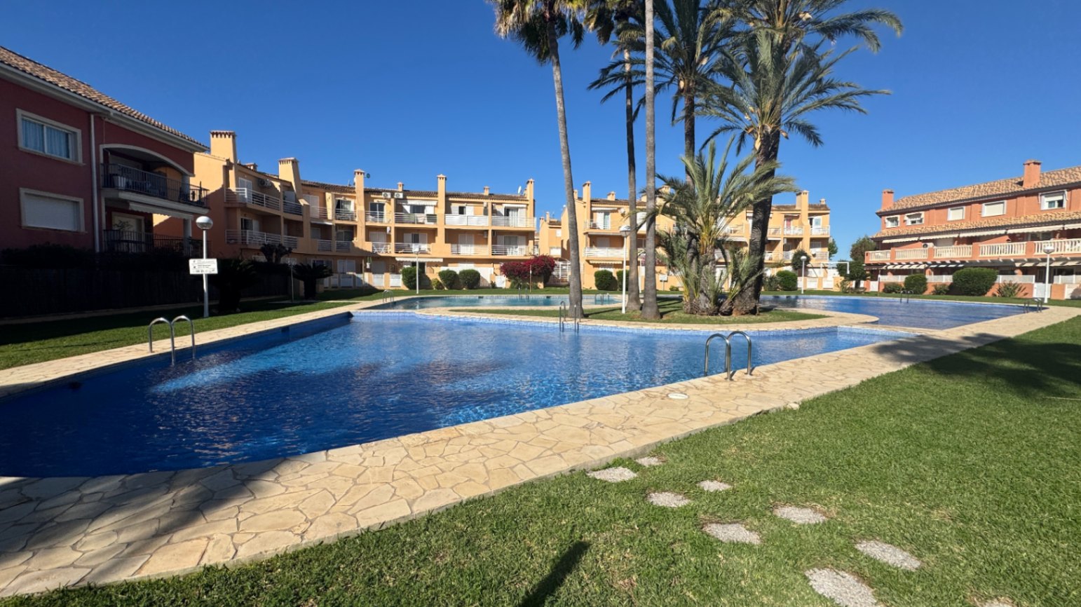 Großer Gemeinschaftspool mit blauen Fliesen, umgeben von einer Steinterrasse, gepflegten Rasenflächen und Palmen in einer mediterranen Wohnanlage.