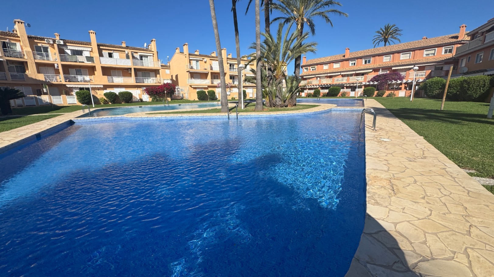 Großer Gemeinschaftspool mit blauen Mosaikfliesen und Natursteinumrandung, eingebettet in eine gepflegte Gartenanlage mit Palmen und Wohngebäuden.