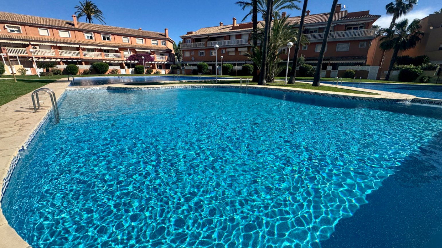 Großer Gemeinschaftspool mit blauen Mosaikfliesen und Steineinfassung, umgeben von gepflegten Grünanlagen und mediterranen Wohngebäuden.