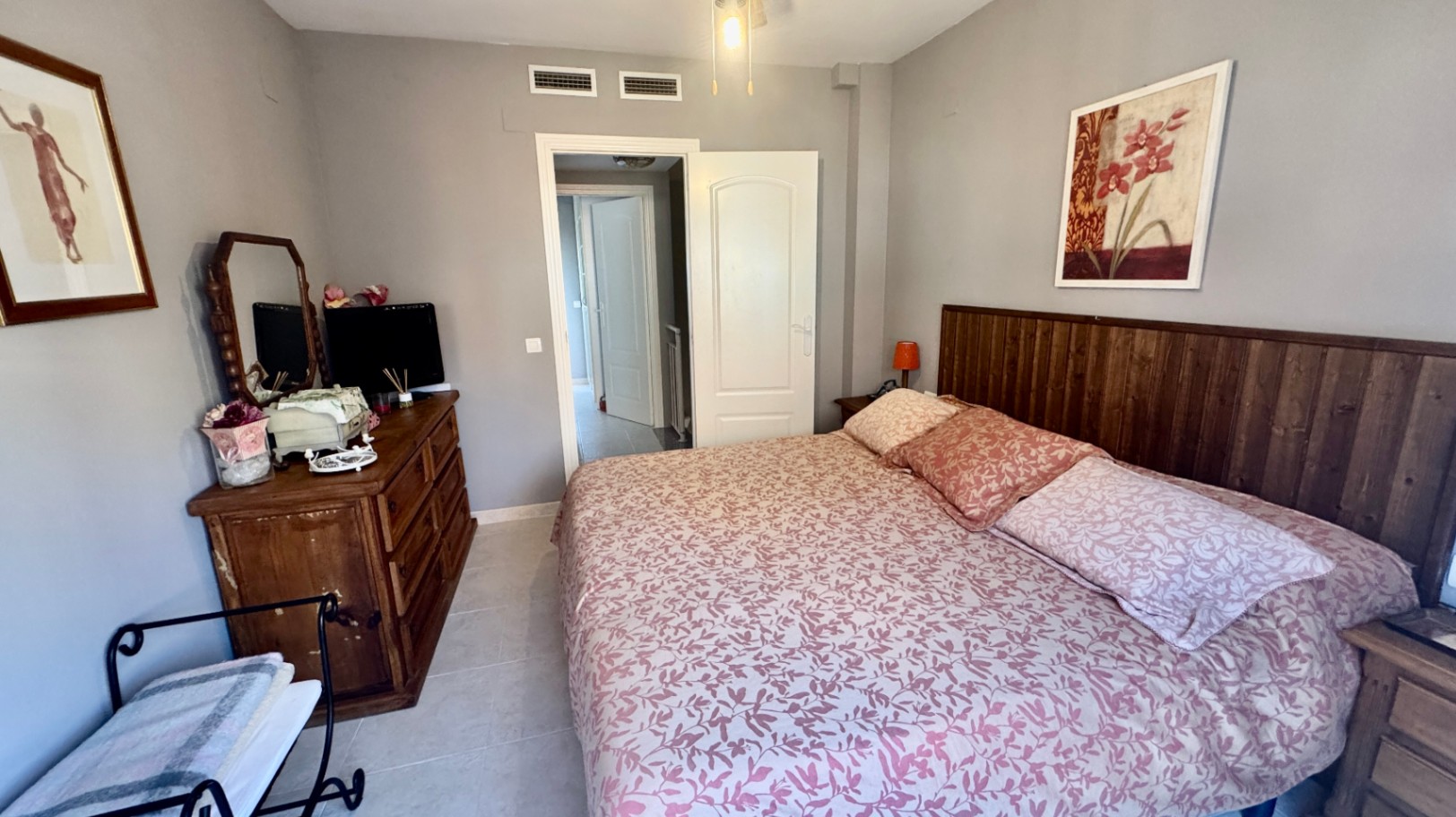 Schlafzimmer mit dunklem Holz-Kopfteil, rustikaler Kommode mit Spiegel und hellgrauen Wänden. Ausgestattet mit Fliesenboden und integrierten Klimaschlitzen.