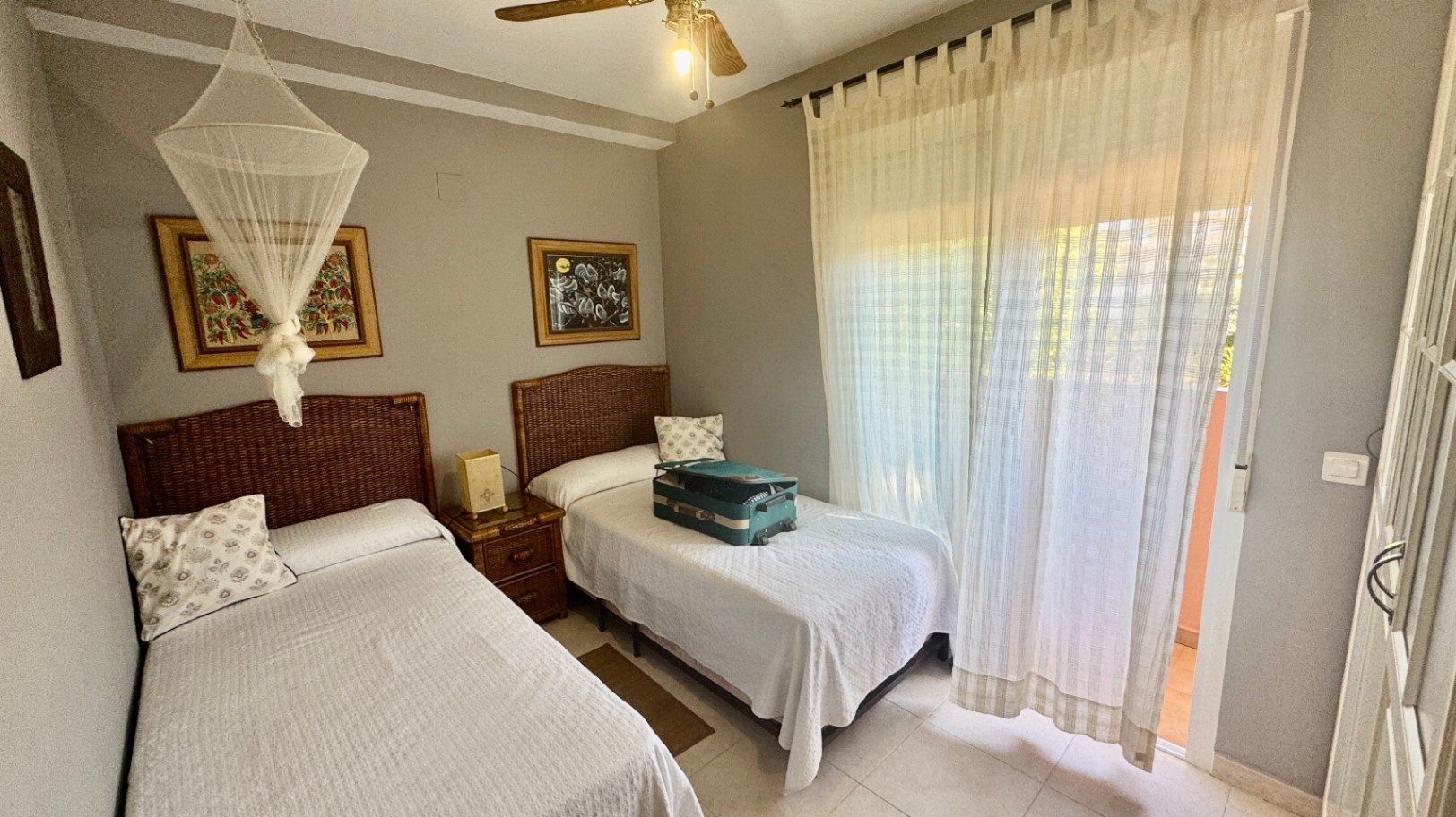 Schlafzimmer mit zwei Einzelbetten, Kopfteilen aus Korbgeflecht und Zugang zum Balkon. Der Raum ist mit einem Deckenventilator und hellen Fliesen ausgestattet.