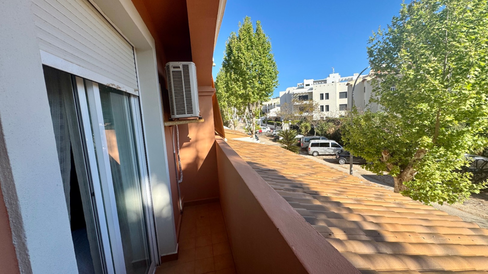 Schmaler Balkon mit Terrakotta-Fliesen, Außeneinheit der Klimaanlage und Blick auf eine von Bäumen gesäumte Straße mit Wohngebäuden.