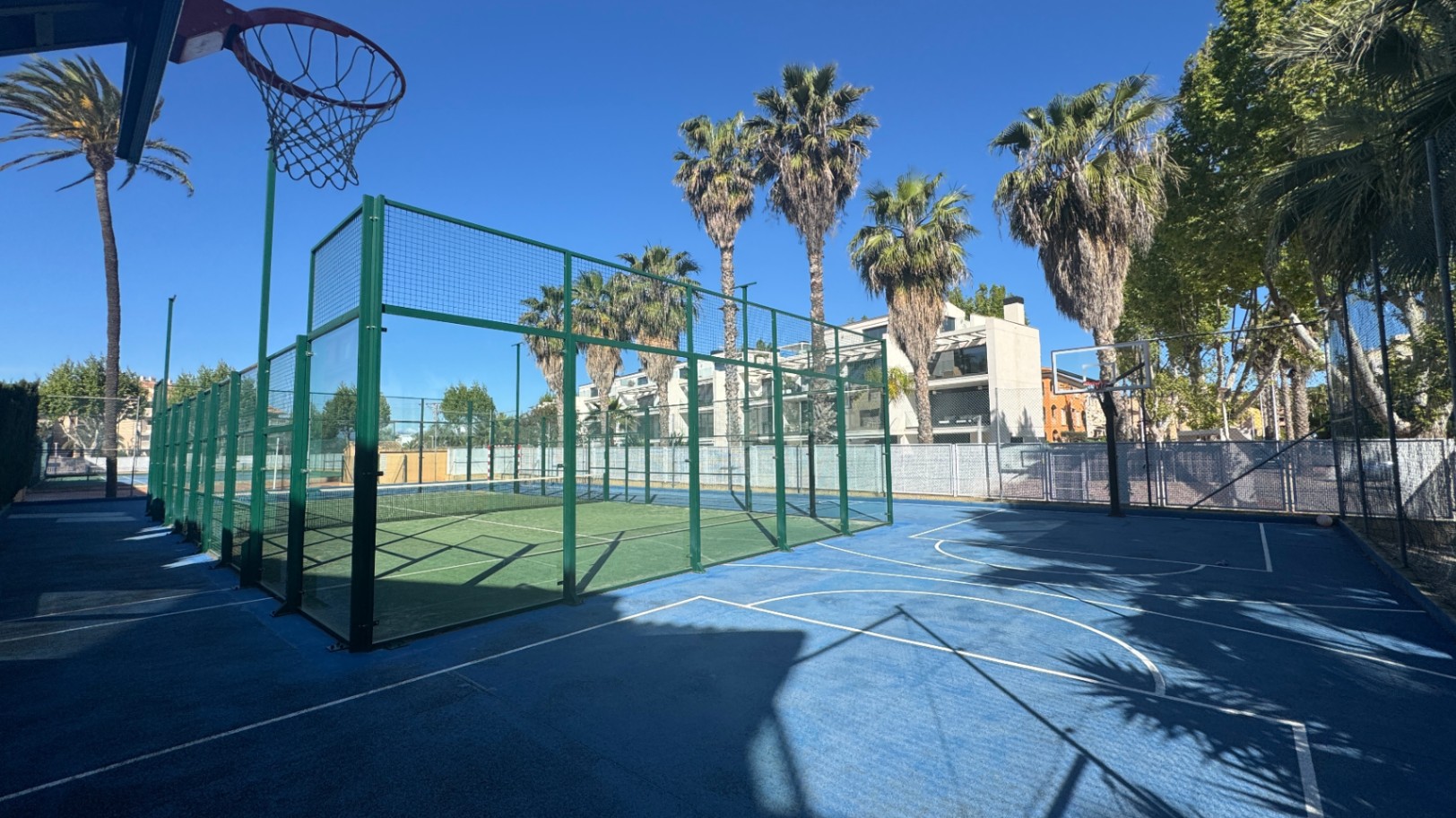 Sportbereich im Freien mit einem grünen Padel-Tennisplatz mit Glaswänden und einem angrenzenden blauen Basketballplatz, umgeben von Palmen.
