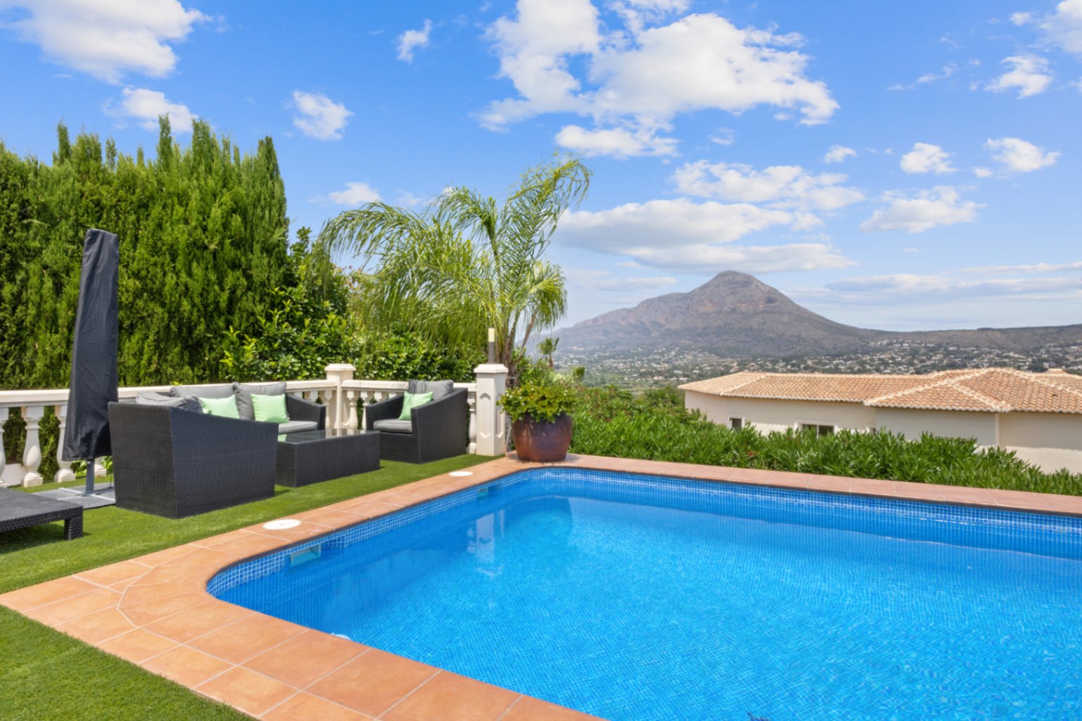 Außenpool mit Terrakotta-Einfassung, Lounge-Bereich auf Kunstrasen mit Polyrattan-Möbeln und Panoramablick auf die umliegende Berglandschaft.