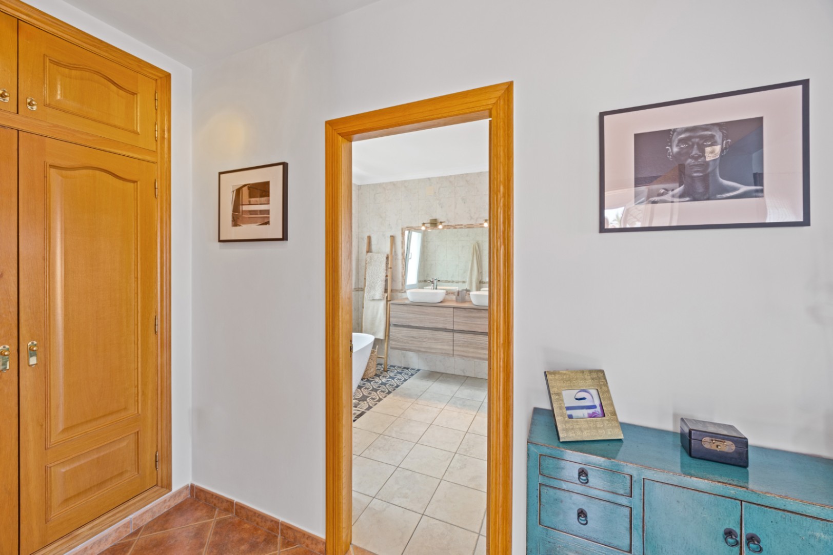 Blick vom Flur mit Einbauschränken aus Holz und blauem Sideboard in ein Badezimmer mit Doppelwaschtisch, Holzunterschrank und hellen Fliesen.