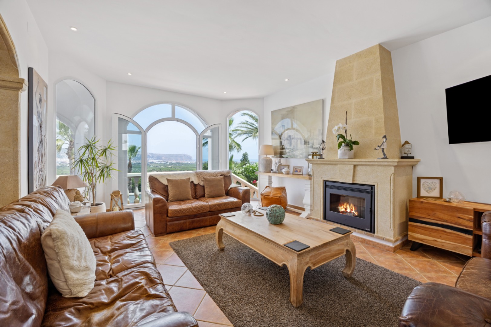 Elegantes Wohnzimmer mit Steinkamin, Ledersofas und Rundbogentüren, die auf eine Terrasse mit Panoramablick auf das Meer und die Berge führen.