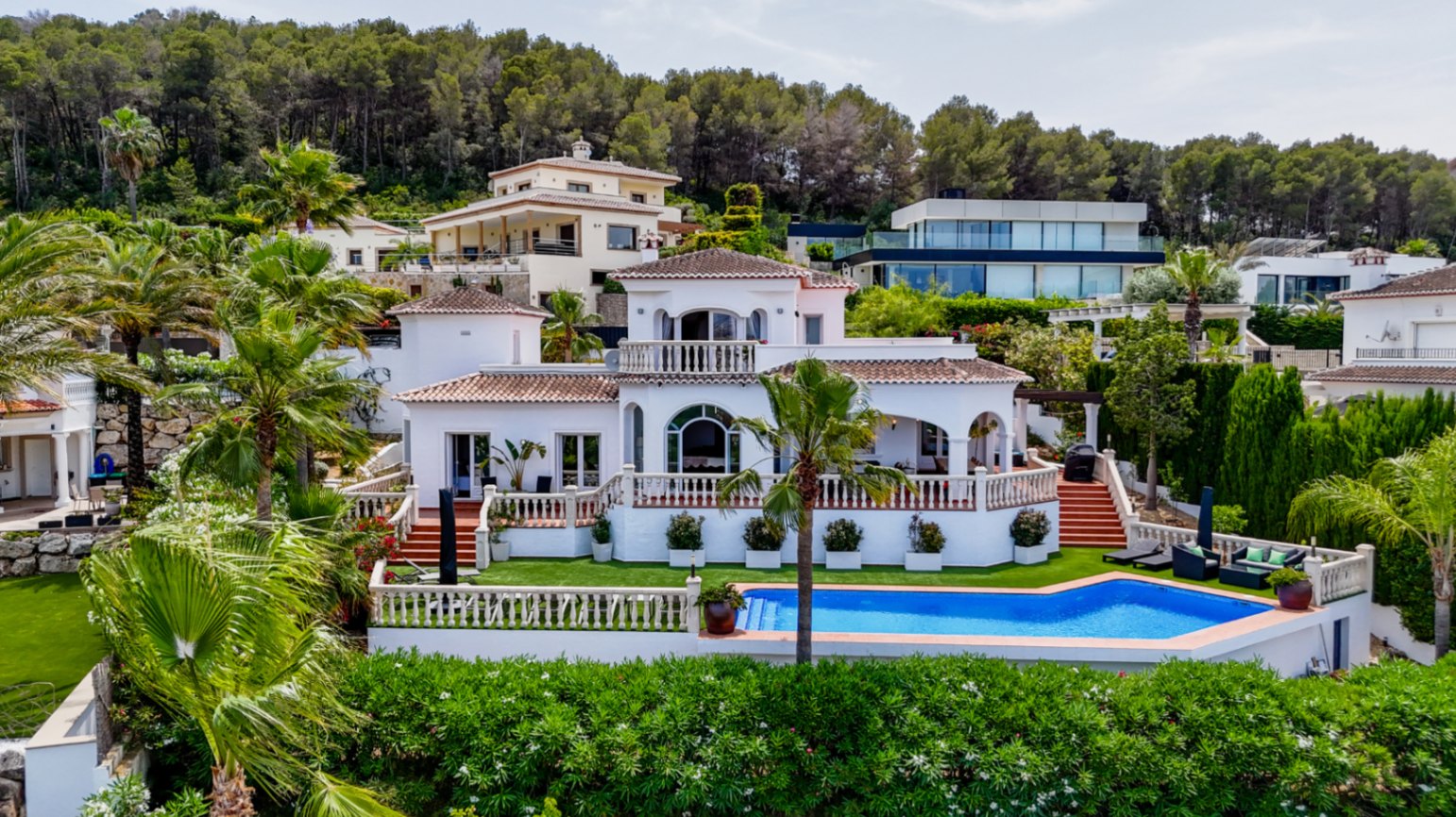 Erhöhte Ansicht einer weißen mediterranen Villa mit Terrakottadächern, Rundbogenfenstern und privatem Pool, umgeben von Palmen und Pinienwald.
