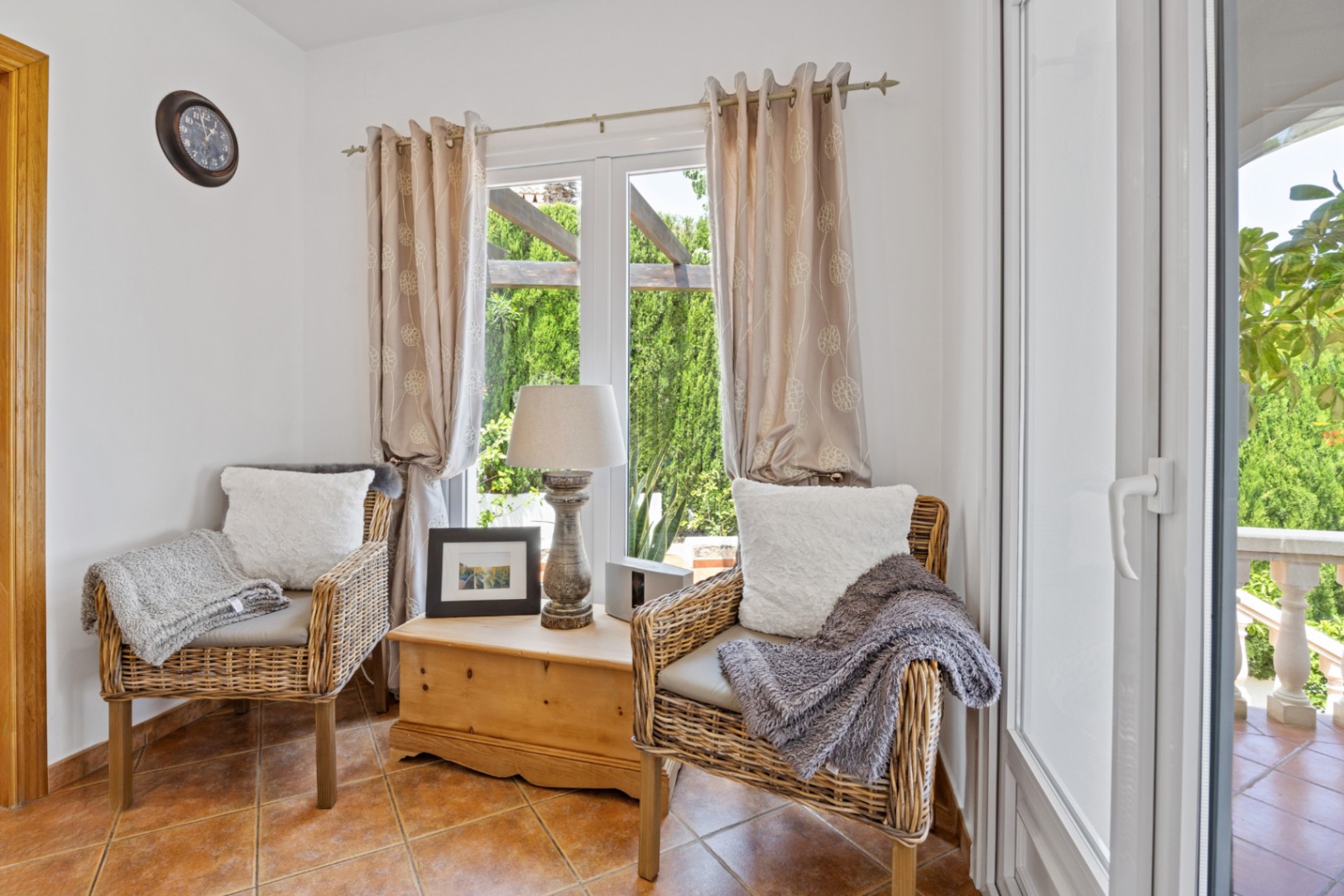 Gemütliche Sitzecke mit zwei Korbsesseln, einer rustikalen Holztruhe und Terrakottaboden. Große Fenster ermöglichen den Blick in den Garten und auf die Pergola.
