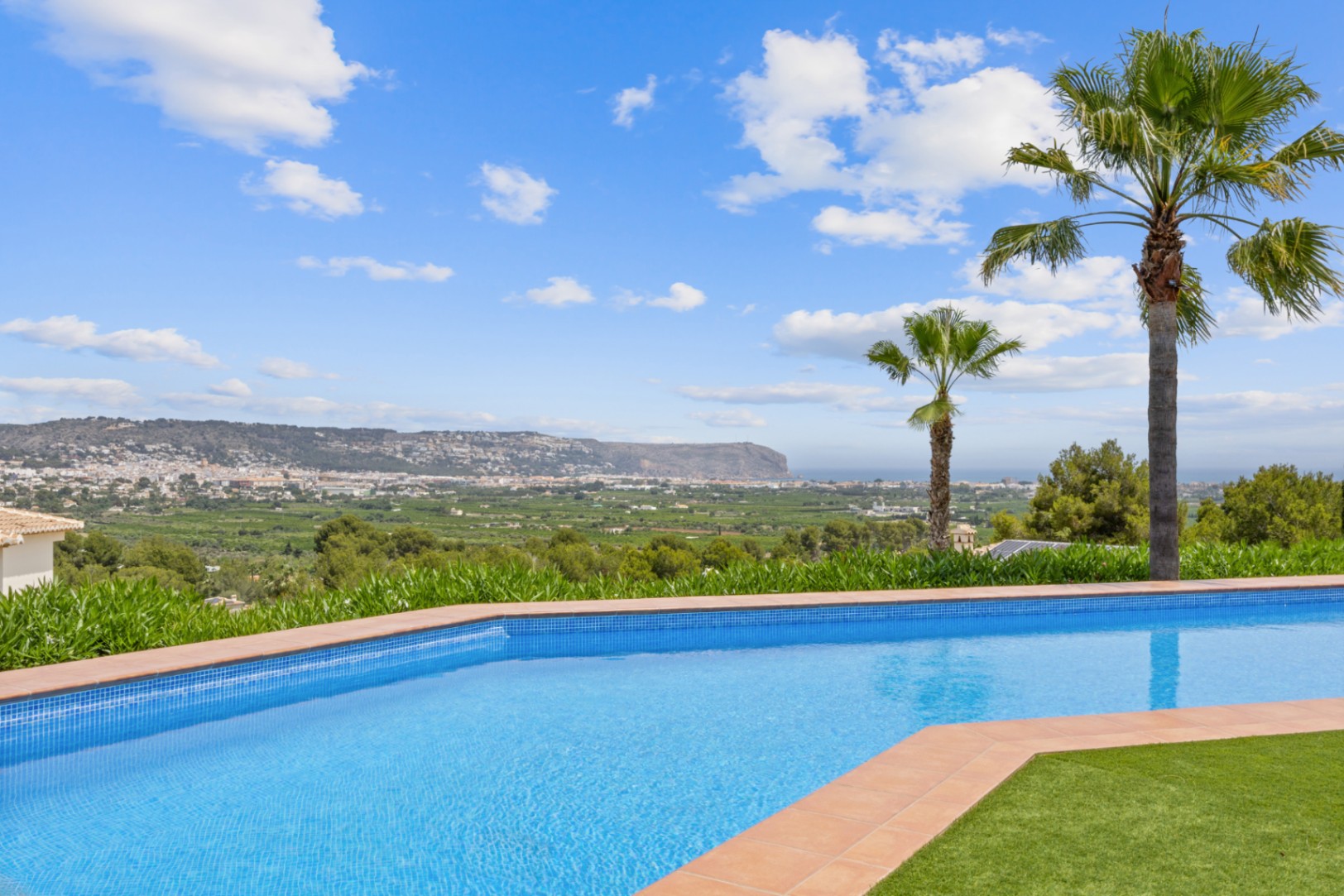 Geschwungener Pool mit blauen Mosaikfliesen und Terrakotta-Einfassung, mit weitem Panoramablick über das Tal und die Küstenstadt.