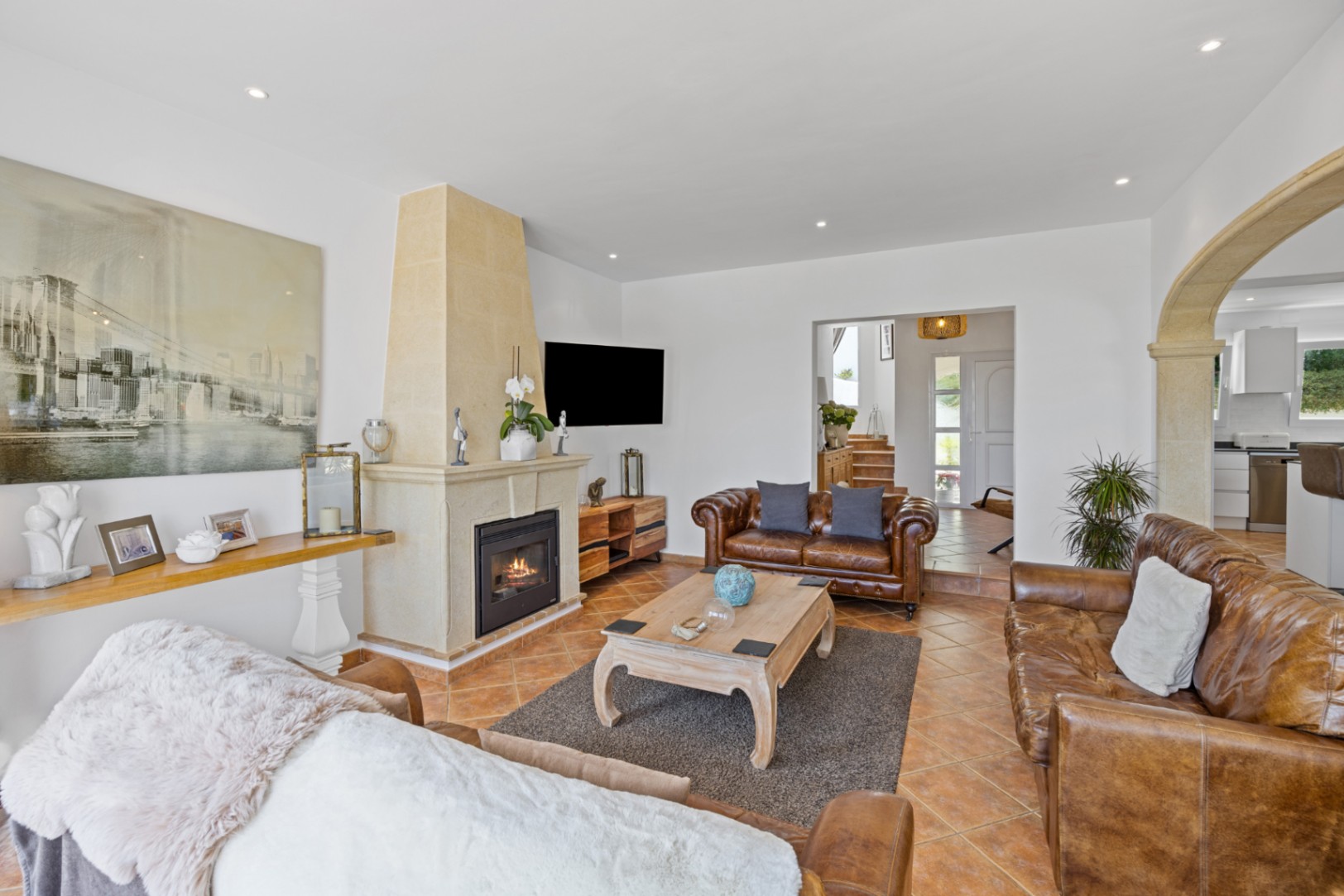 Großzügiges Wohnzimmer mit braunen Ledersofas, einem Natursteinkamin und Terrakottaböden. Ein steinerner Rundbogen führt zur angrenzenden Küche.
