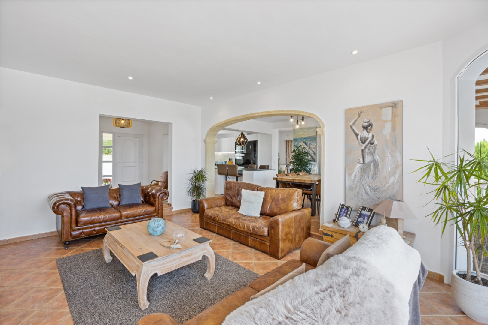 Großzügiges Wohnzimmer mit braunen Ledersofas, Terrakottaboden und einem Steinbogen, der zum Essbereich und der offenen Küche führt.