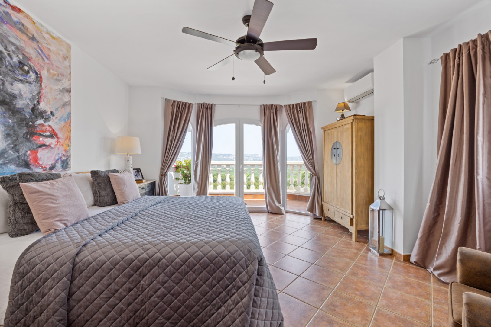 Helles Schlafzimmer mit Terrakottaboden, Deckenventilator und bogenförmigen Glastüren, die auf einen Balkon mit weitem Landschaftsblick führen.