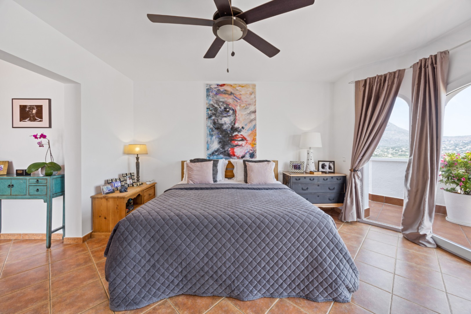 Helles Schlafzimmer mit Terrakottafliesen, Deckenventilator und Glasschiebetüren, die auf einen privaten Balkon mit Panoramablick auf die Berge führen.