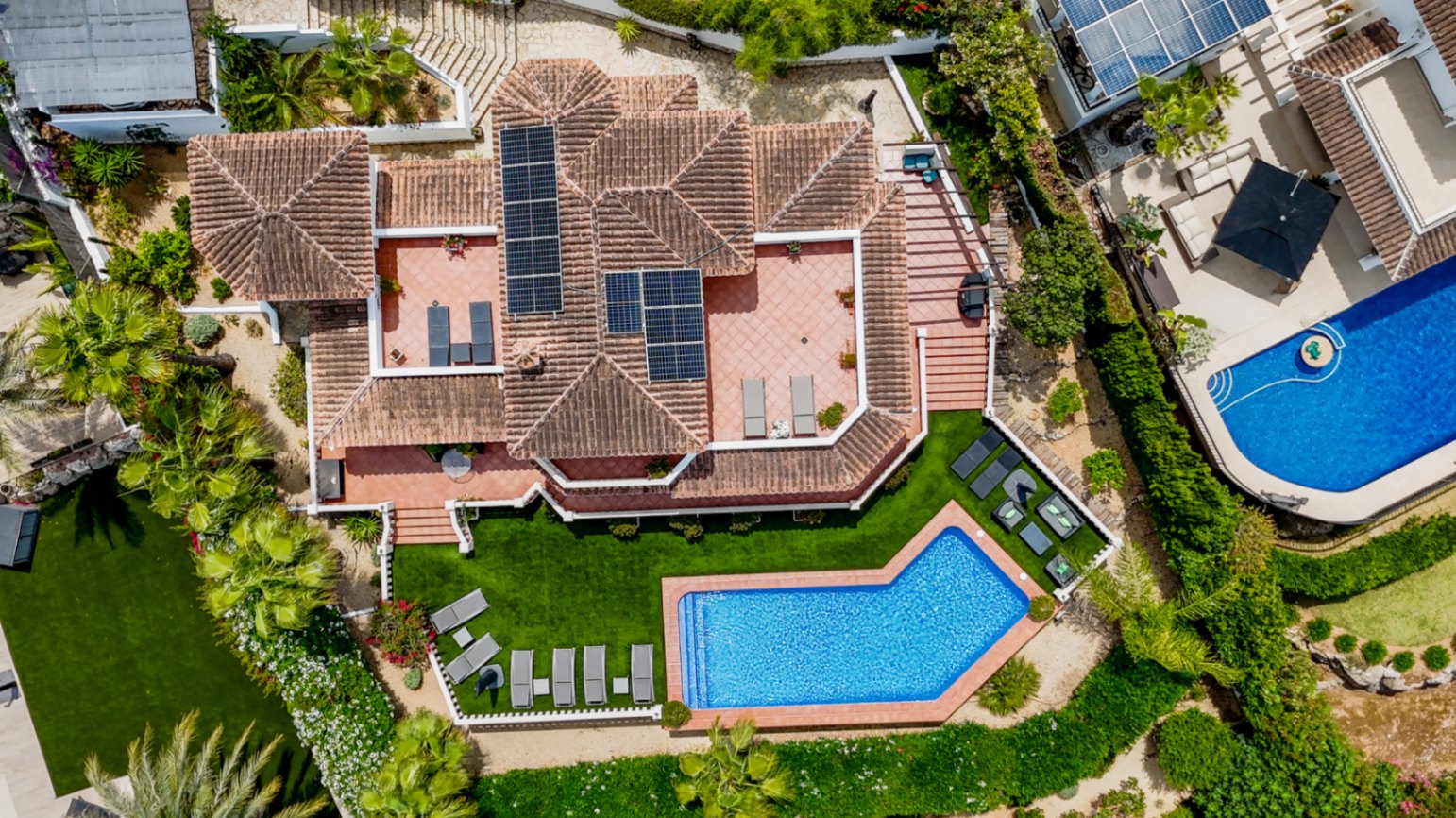 Luftaufnahme einer mediterranen Villa mit Terrakottadach, Solarmodulen, mehreren Sonnenterrassen und einem eckigen Pool inmitten gepflegter Rasenflächen.