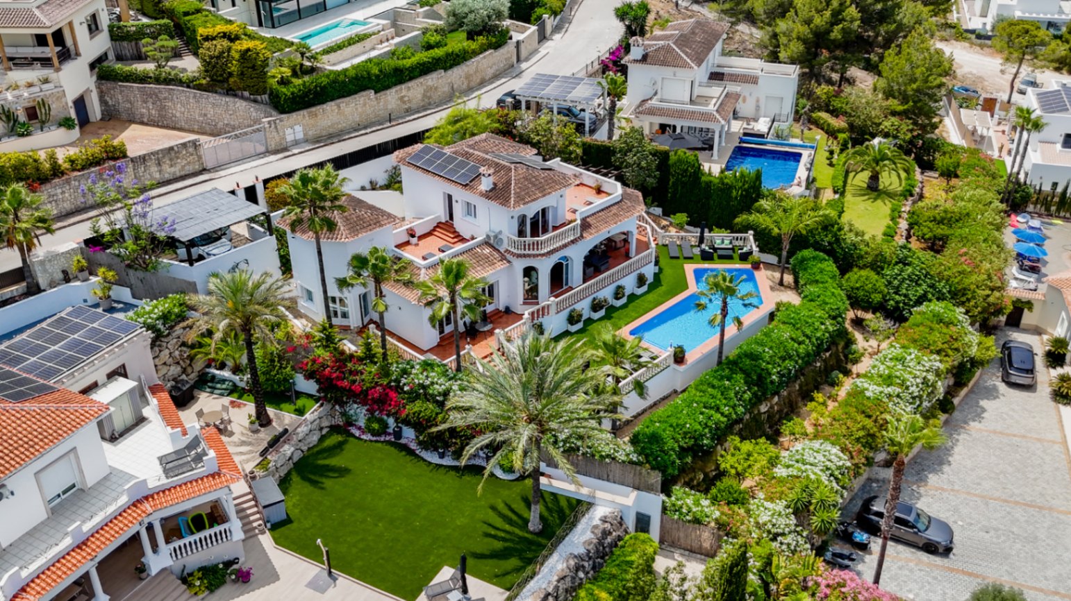 Luftaufnahme einer mediterranen Villa mit Terrakottadächern, Sonnenkollektoren, privatem geometrischem Pool und gepflegten Gärten mit Palmen.