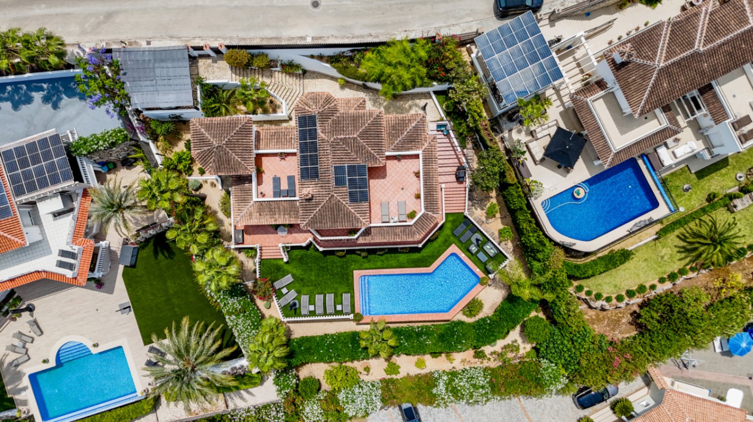 Luftaufnahme einer Villa mit Terrakottadach und Solarmodulen, einem geometrischen Pool und angelegten Gärten mit Palmen und Sonnenterrassen.