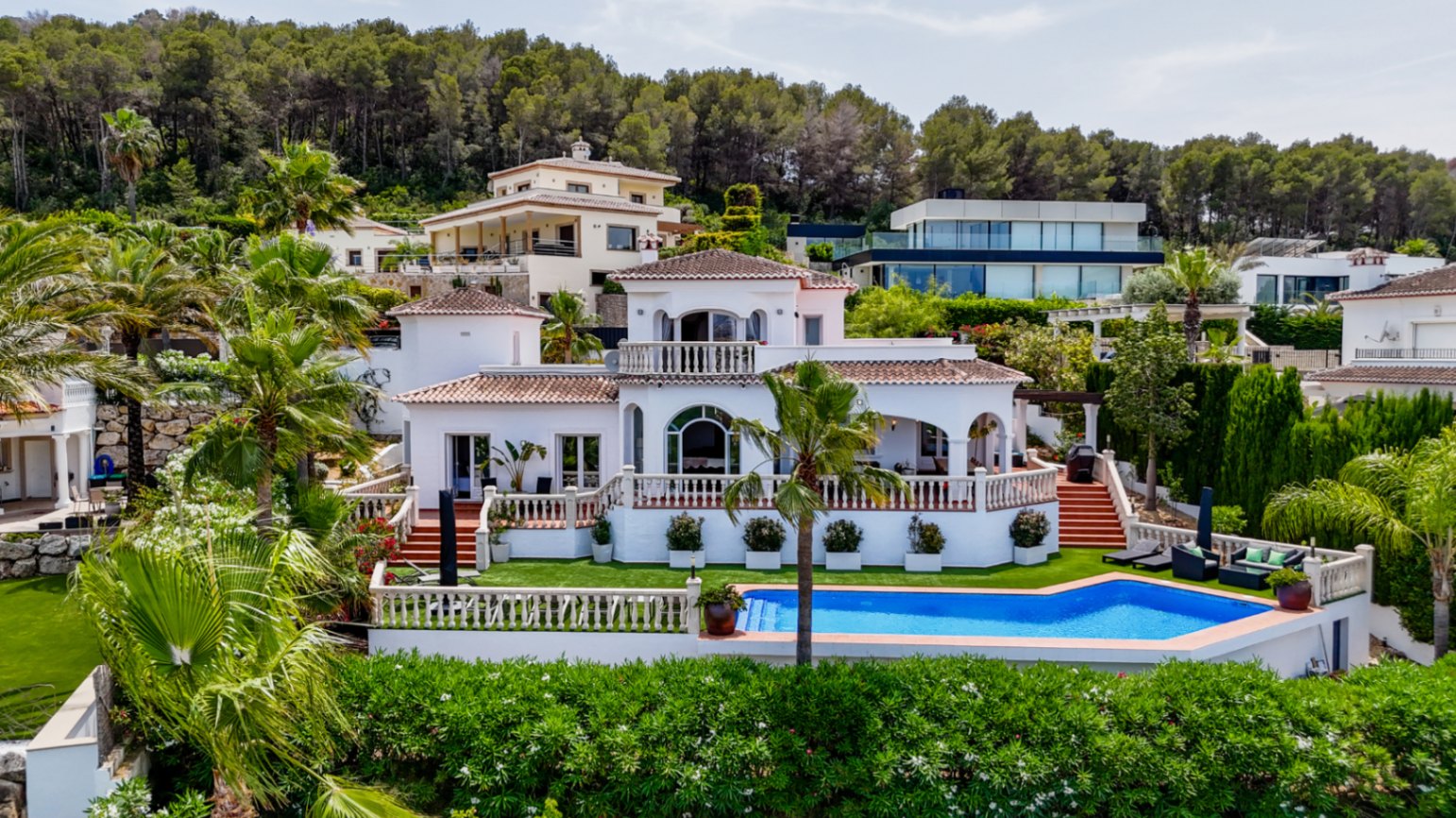 Mediterrane Villa mit weißer Fassade, Terrakottadach, mehreren Terrassenebenen und privatem Pool, umgeben von Palmen und bewaldeten Hügeln.