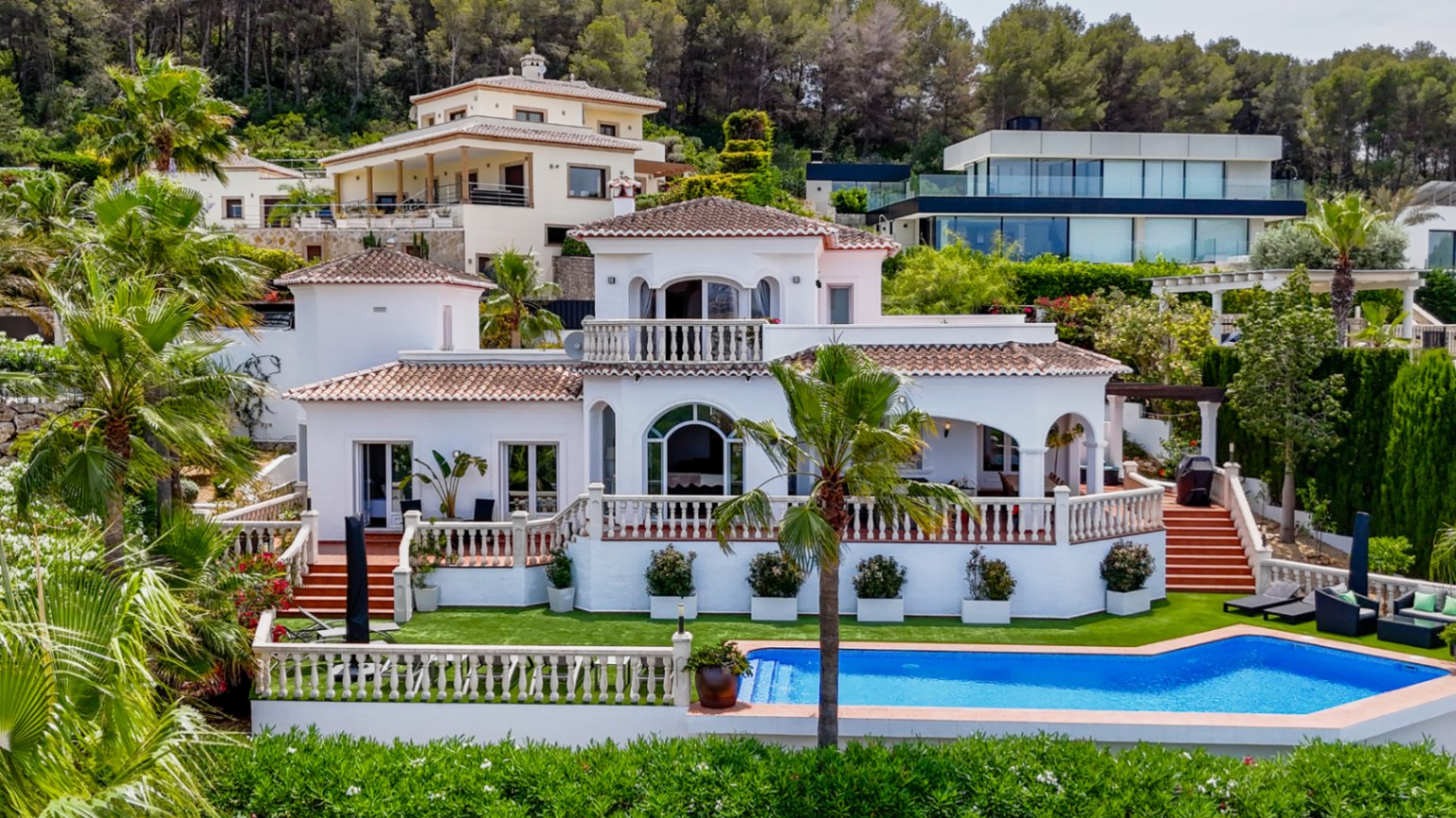Mediterrane Villa mit weißer Fassade, Terrakottadach und abgestuften Terrassen mit Steinbalustraden über einem privaten Pool und Palmengarten.