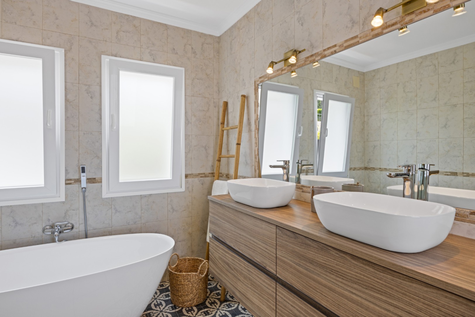 Modernes Badezimmer mit Doppelwaschtisch aus Holz, freistehender Badewanne und gemusterten Bodenfliesen, ergänzt durch warmes Licht und zwei Fenster.