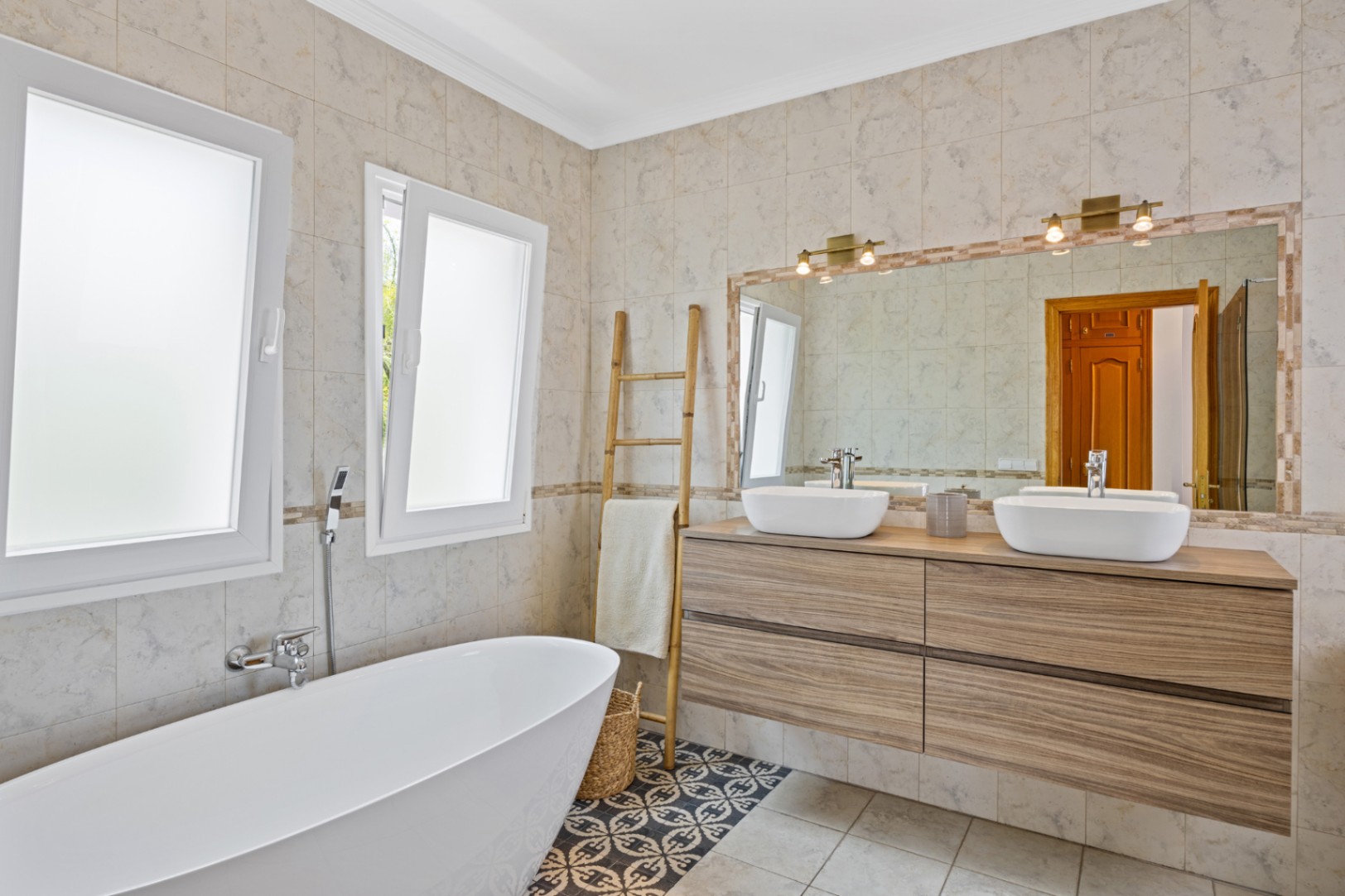 Modernes Badezimmer mit freistehender Wanne, Doppelwaschtisch aus Holz und dekorativen Bodenfliesen. Großer Spiegel mit Messinglampen und mattierten Fenstern.