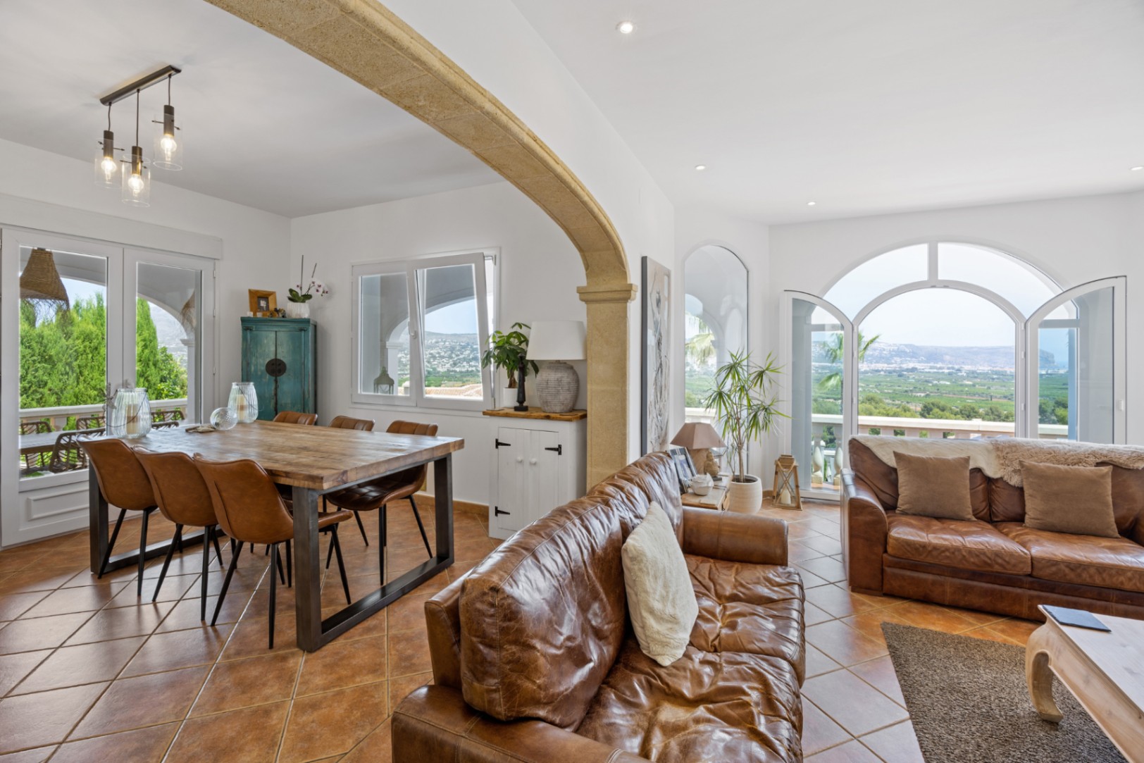 Offener Wohnbereich mit Natursteinbogen, Ledersofas und Holzesstisch. Rundbogen-Glastüren führen zur Terrasse mit weitem Blick über das Tal.
