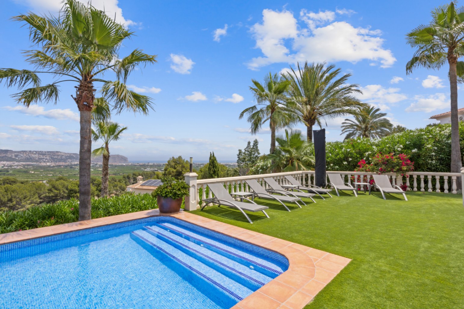 Poolterrasse mit grauen Sonnenliegen auf Kunstrasen, weißer Steinbalustrade und Panoramablick über das Tal bis hin zur Küste.