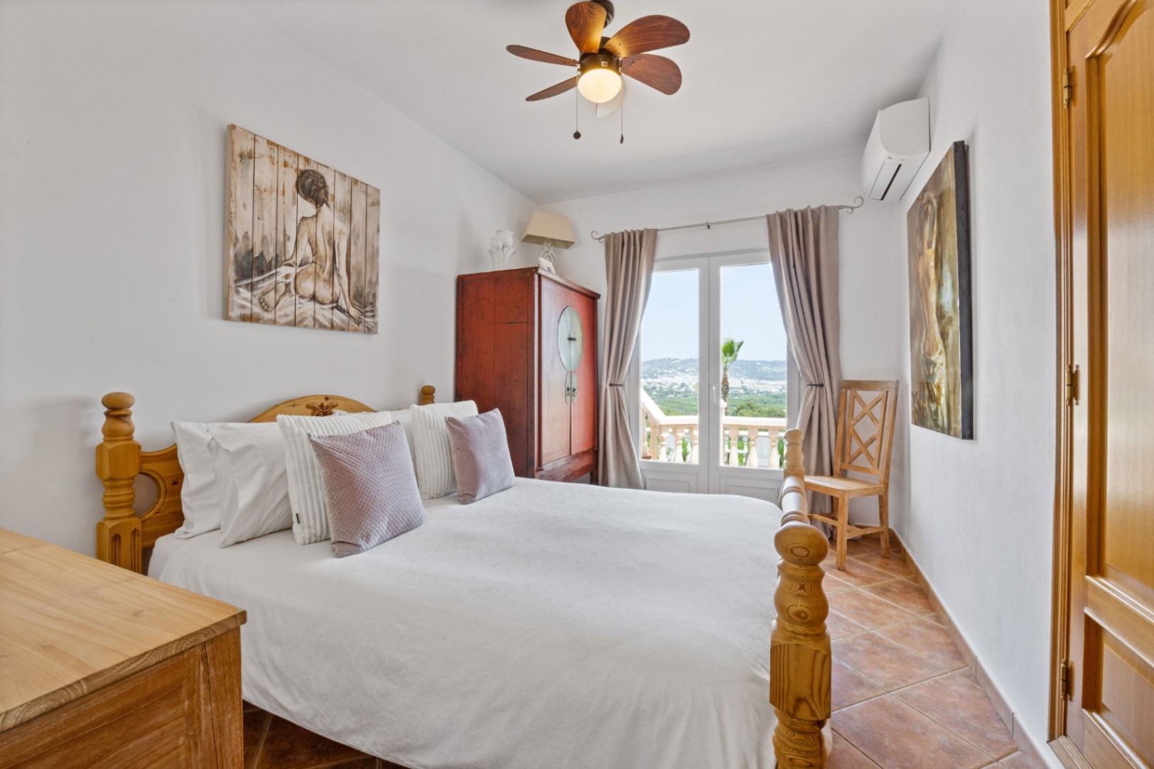 Schlafzimmer mit Holzbett, Terrakottafliesen und Fenstertüren zum Balkon mit Weitblick. Ausgestattet mit Deckenventilator und Klimaanlage.