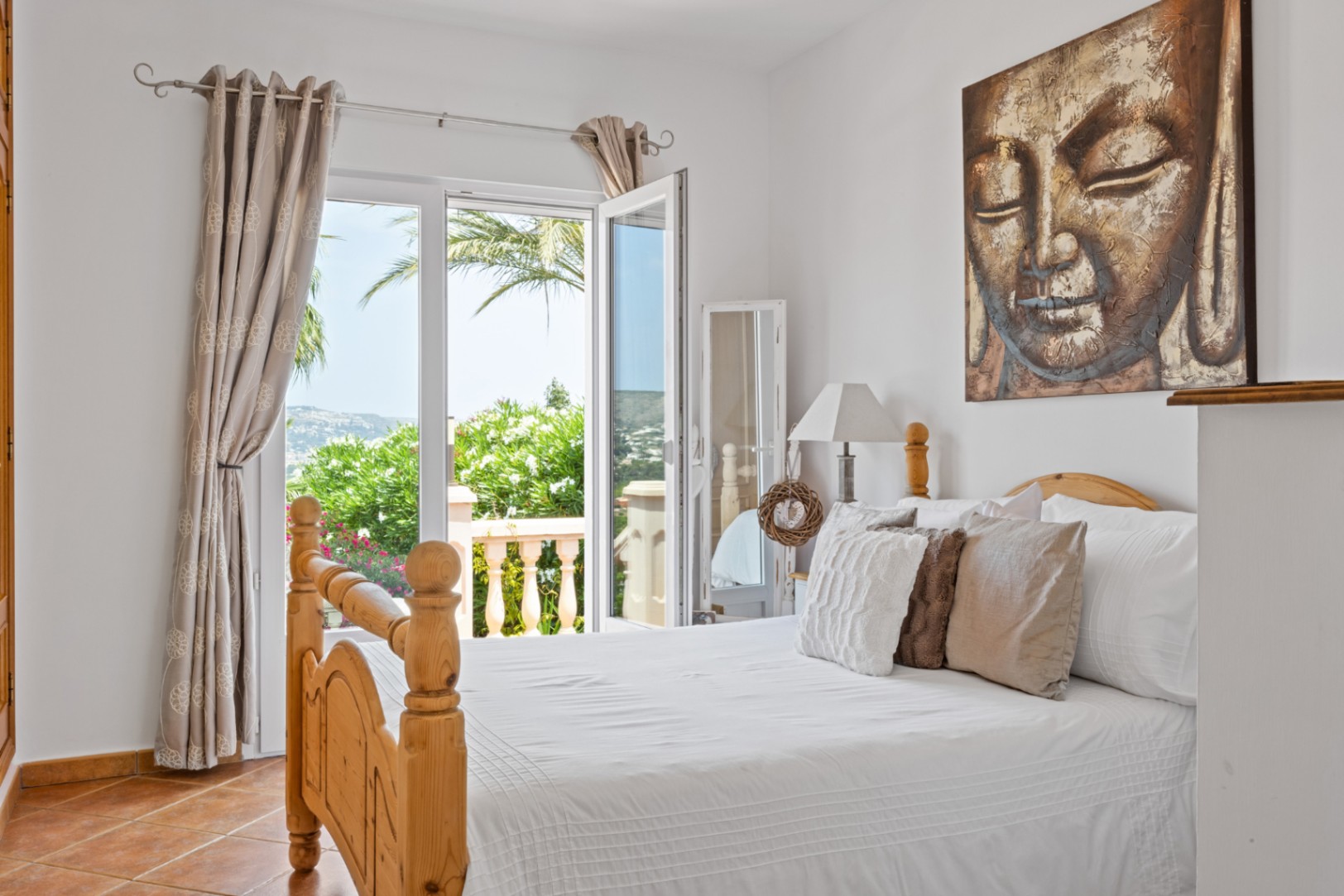 Schlafzimmer mit Holzbettrahmen, Terrakottaboden und großem Buddha-Gemälde. Glastüren führen auf einen Balkon mit Blick auf Palmen und die umliegende Hügellandschaft.