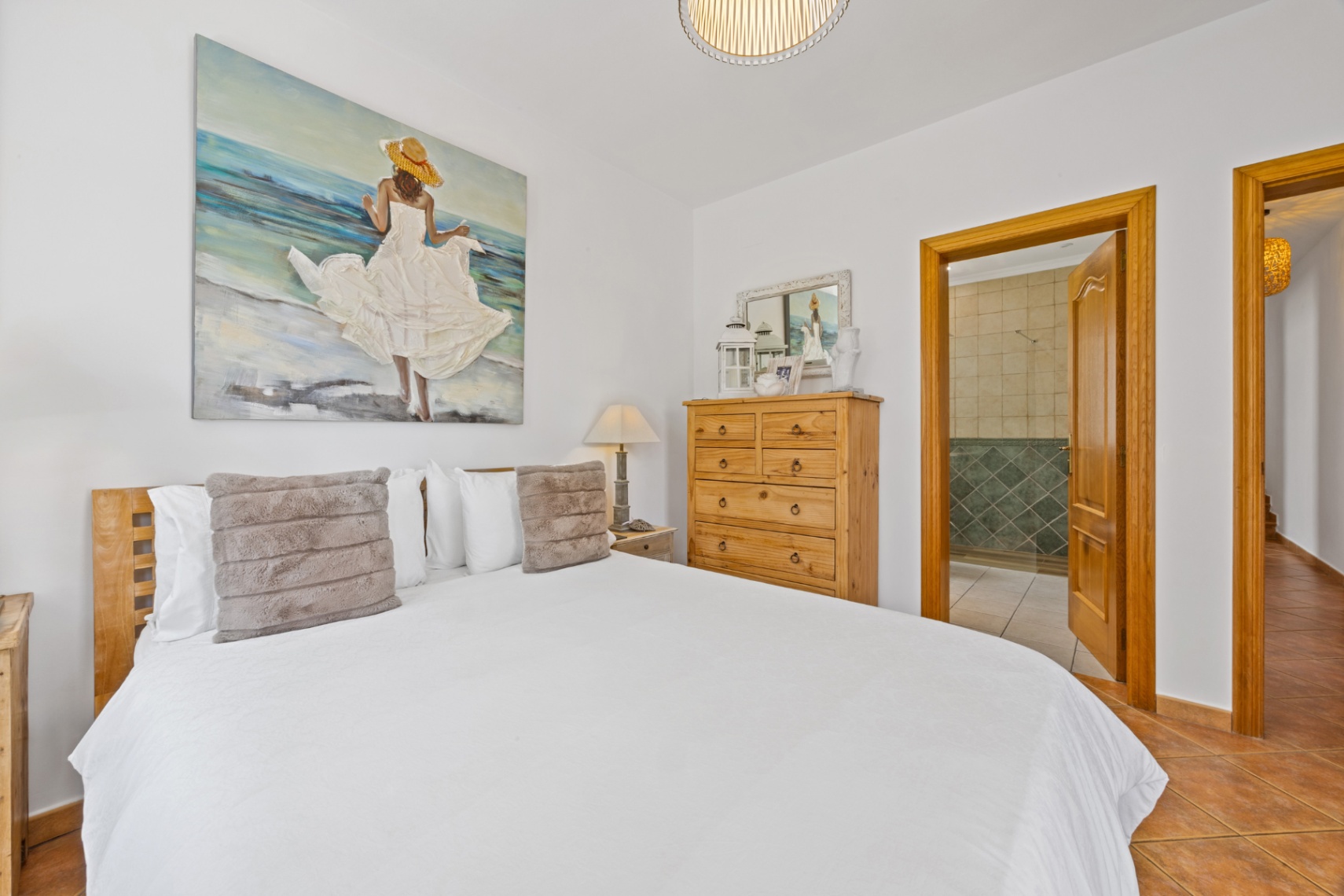 Schlafzimmer mit hölzernem Kopfteil, großem Wandbild und Terrakotta-Fliesenboden. Eine offene Tür führt zum angrenzenden Badezimmer mit grünen Fliesen.