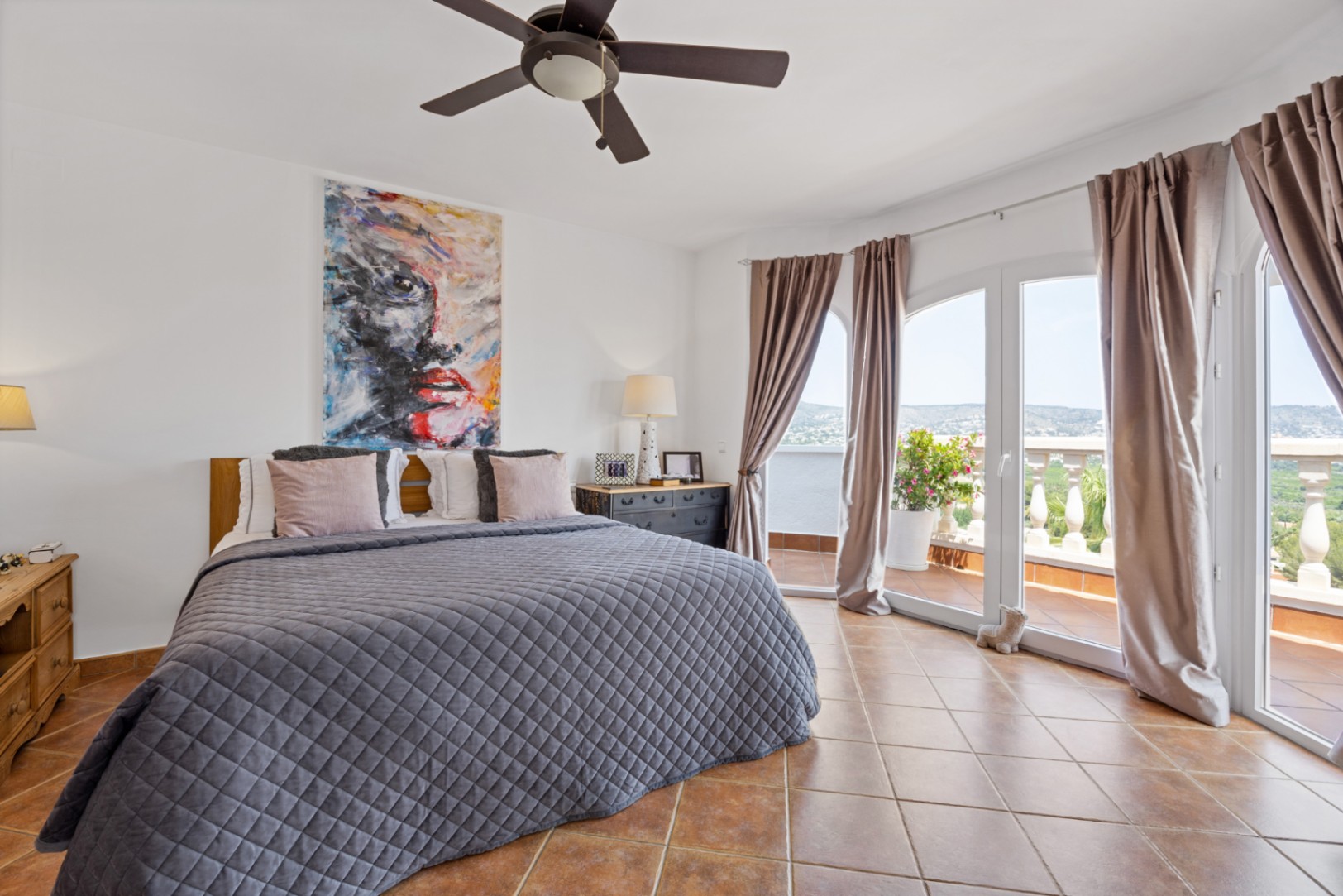 Schlafzimmer mit Terrakottafliesen, Deckenventilator und bogenförmigen Glastüren, die auf eine Terrasse mit weitem Landschaftsblick führen.