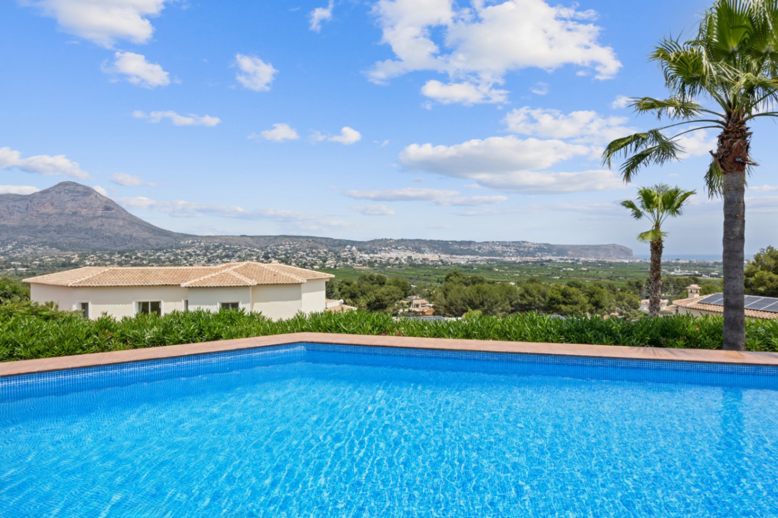 Schwimmbecken mit blauen Mosaikfliesen und Terrakotta-Rand, mit Panoramablick auf das Tal und die Berge unter blauem Himmel.