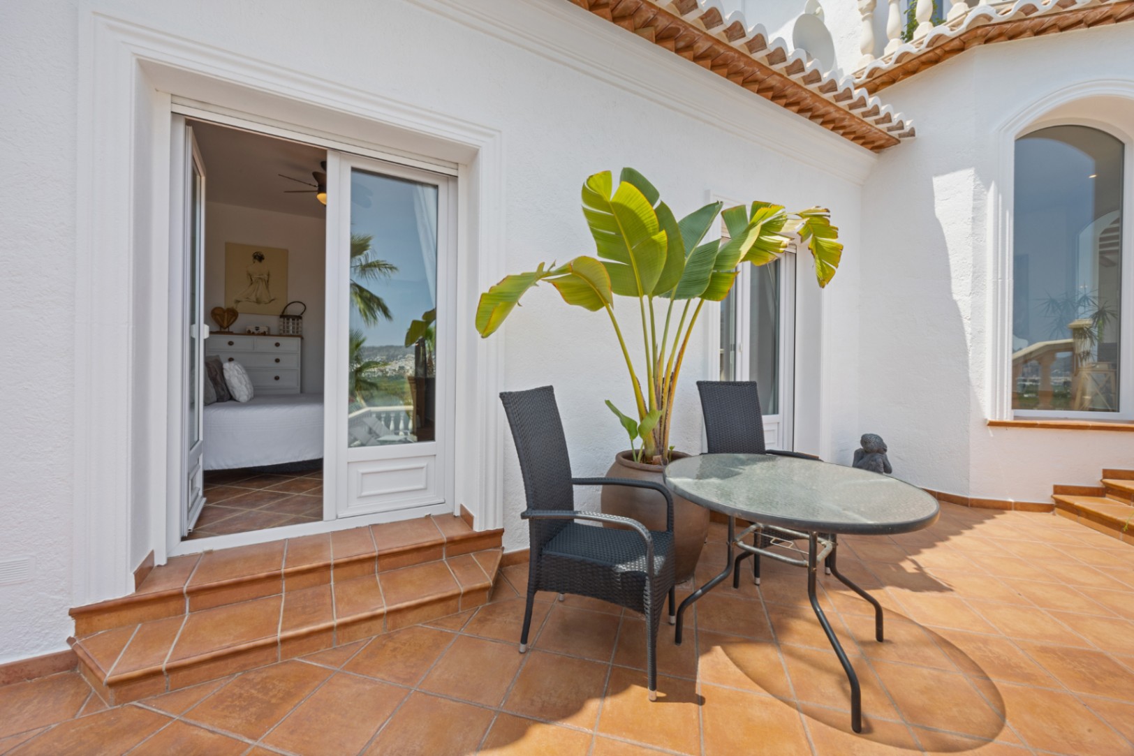 Terrasse mit Terrakottafliesen, Außenmöbeln und direktem Zugang zu einem hellen Schlafzimmer durch große Glasschiebetüren.