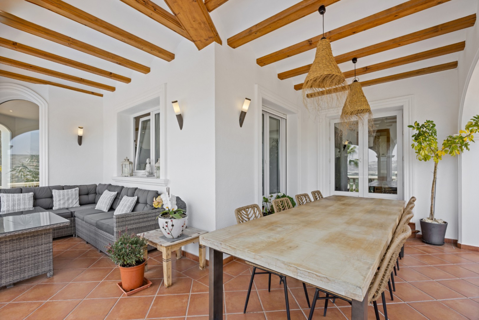 Überdachte Terrasse mit Holzbalkendecke, Terrakottaboden, großem Holzesstisch mit Korbstühlen und einer Lounge-Ecke mit grauem Rattansofa.
