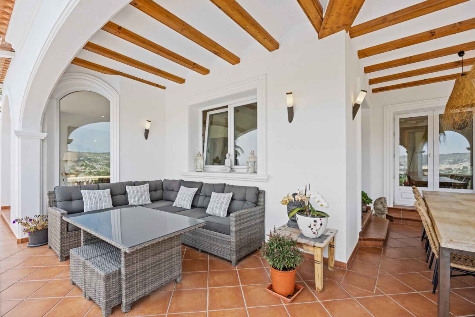 Überdachte Terrasse mit markanten Holzbalken, Terrakottafliesen und einer grauen Rattan-Sitzgruppe. Wandleuchten und ein angrenzender Essbereich ergänzen den Außenraum.