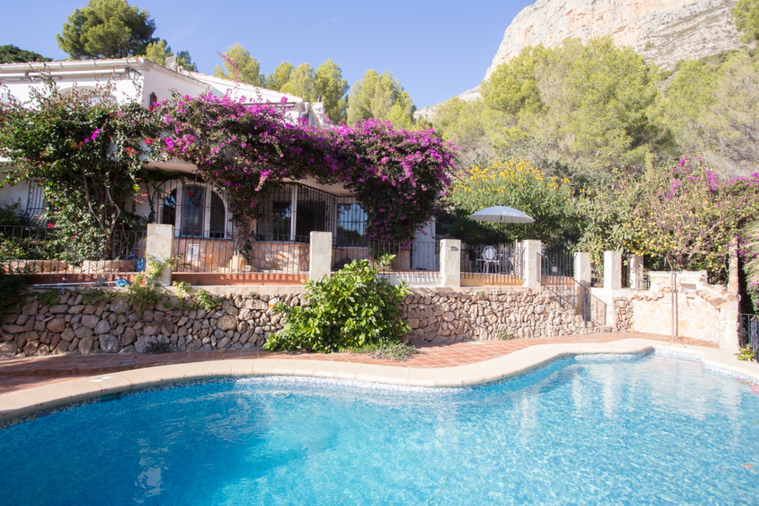 Außenansicht mit geschwungenem Pool, Terrakotta-Terrasse und einer weißen Villa mit violetter Bougainvillea vor einer markanten Felskulisse.